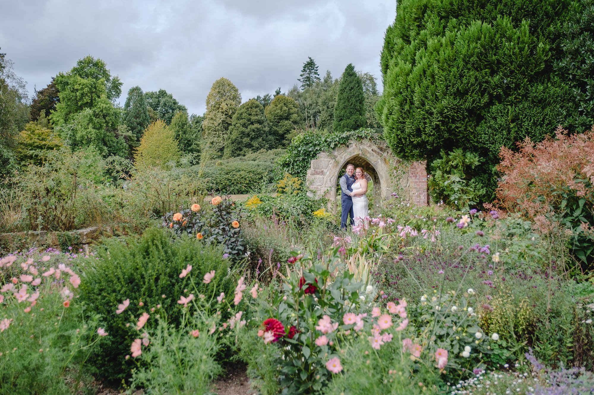 A bride and groom standing together in the lush garden at Sctoney Castle with a stone archway behind them, surrounded by various colorful flowers and green trees.