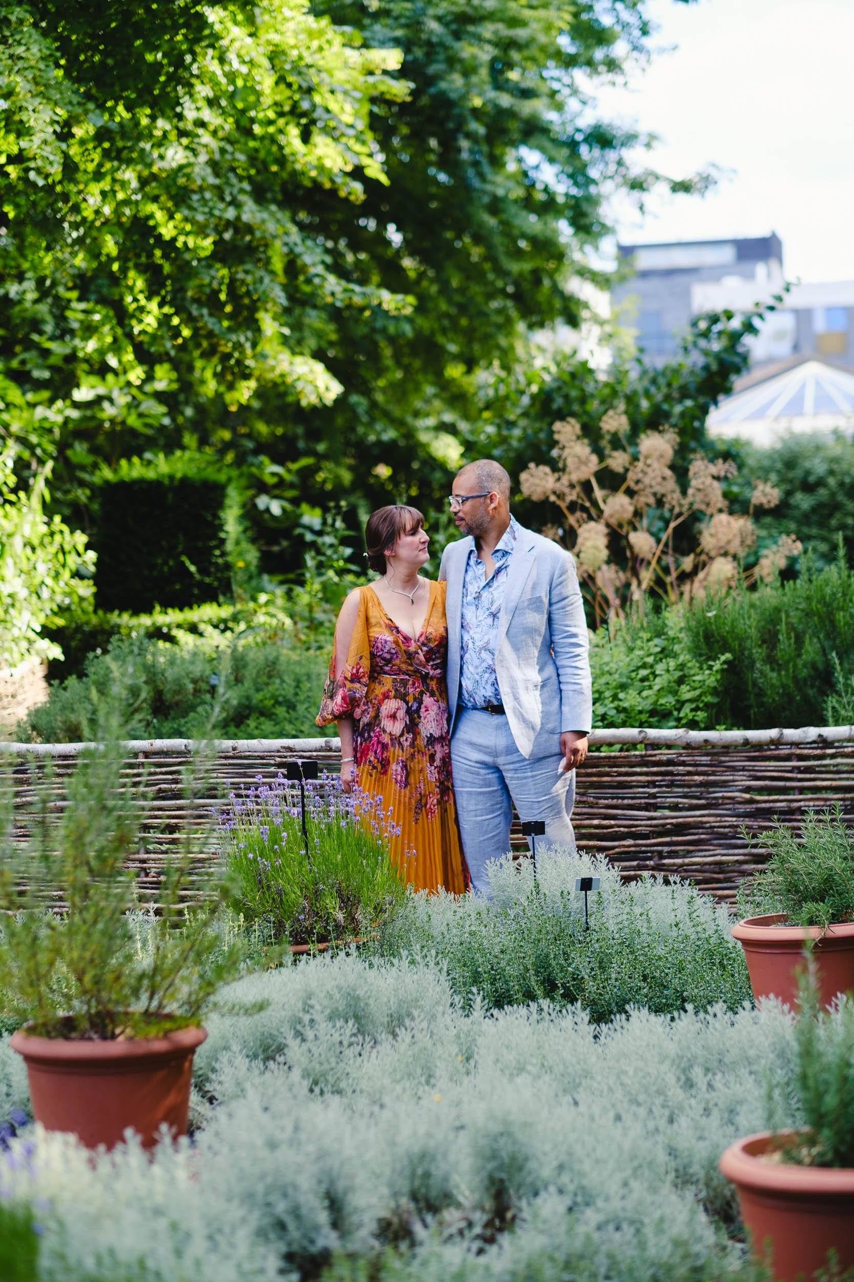 A couple standing together in the garden of The Museum, of the Home, London,, surrounded by various plants and flowers, with a cityscape in the background.