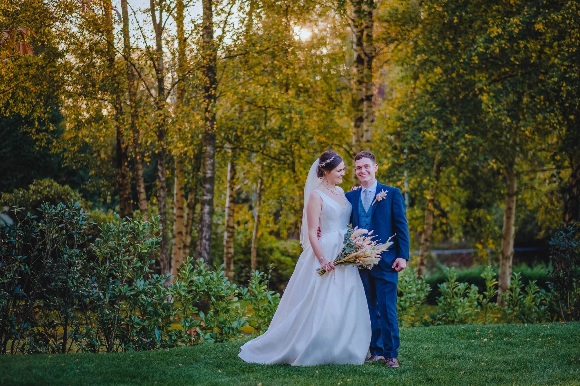 A bride and groom smiling and posing together outdoors in a park surrounded by autumn trees. The bride is wearing a white wedding gown and holding a bouquet of wildflowers. The groom is dressed in a blue suit with a light blue tie and boutonniere.