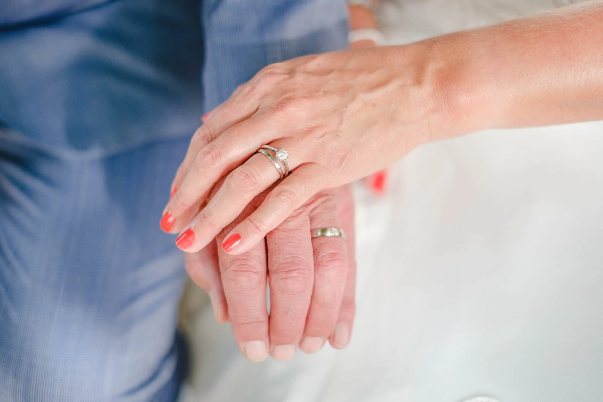 Close-up of a couple's hands showing wedding rings, the woman's hand is resting on the man's hand on a white fabric surface.