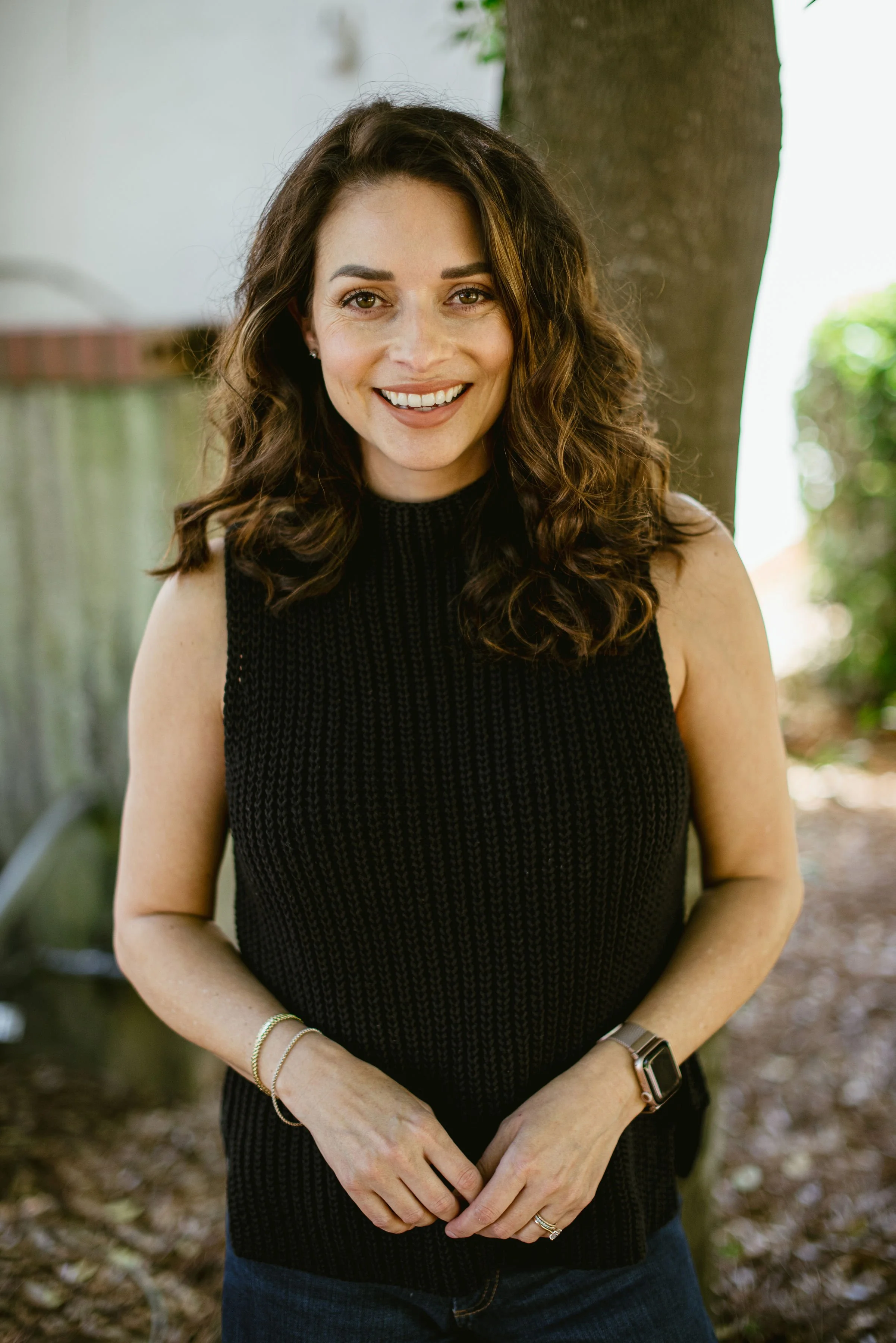 Beth Dent black sleeveless turtleneck standing outside in front of tree trunk brunette wavy hair