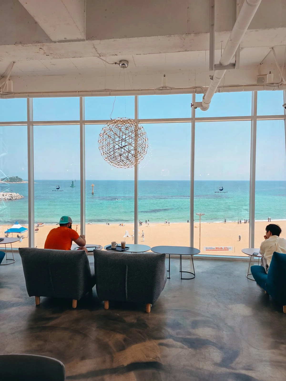 Inside a cafe where two men are sitting at tables and facing away from the camera looking out of floor to ceiling windows that show a view of a sandy beach and turquoise blue water on a sunny day