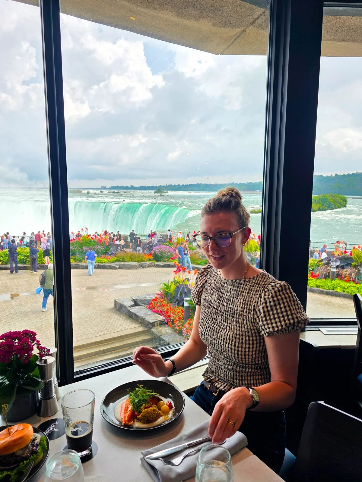 Helena picking up a fork at a restaurant table about to eat a meal with a view of Niagara Falls Horseshoe Falls behind her out the window