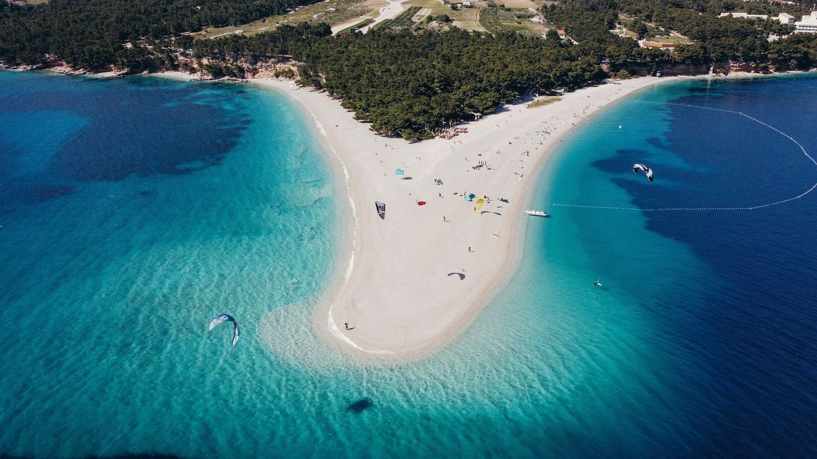 A drone photo looking down on a white sand bar beach surrounded by turquoise blue water. People can be seen on the beach and windsurfers in the water