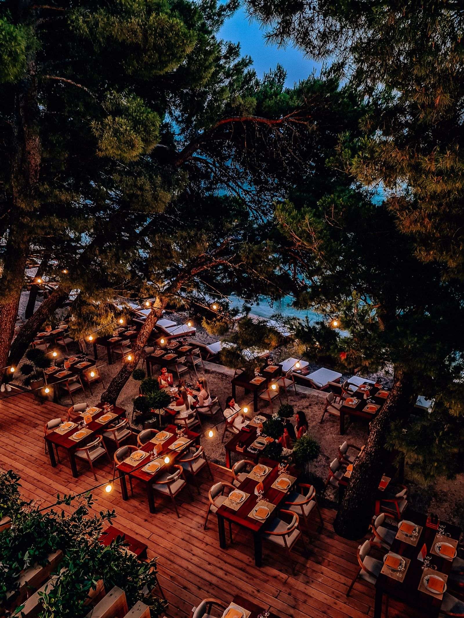 looking down on a dining area set up on the beach next to the sea below the trees with fairy lights strung around