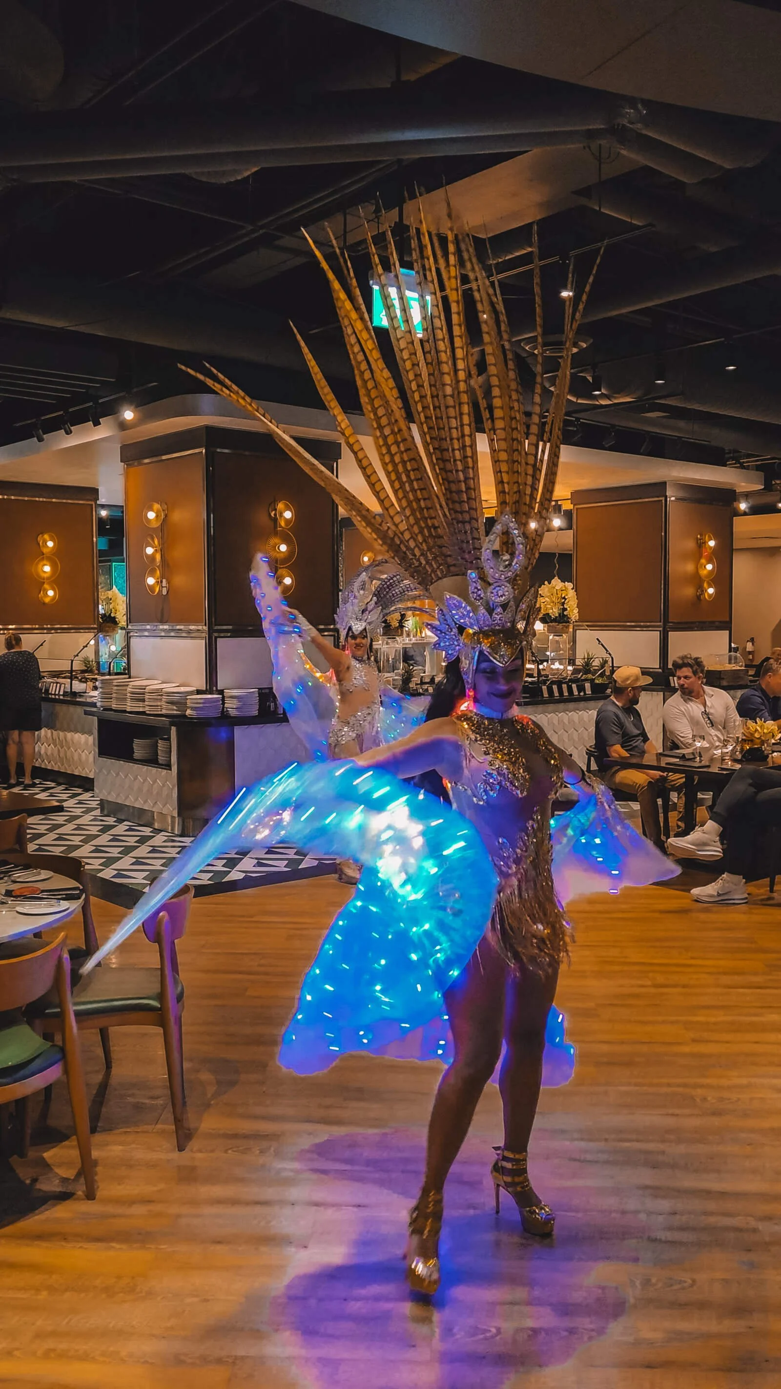 A woman dressed in a golf salsa outfit with a feather crown on her head and a cape that is lit up with blue lights. She is dancing in a Brazilian Steakhouse restaurant performance