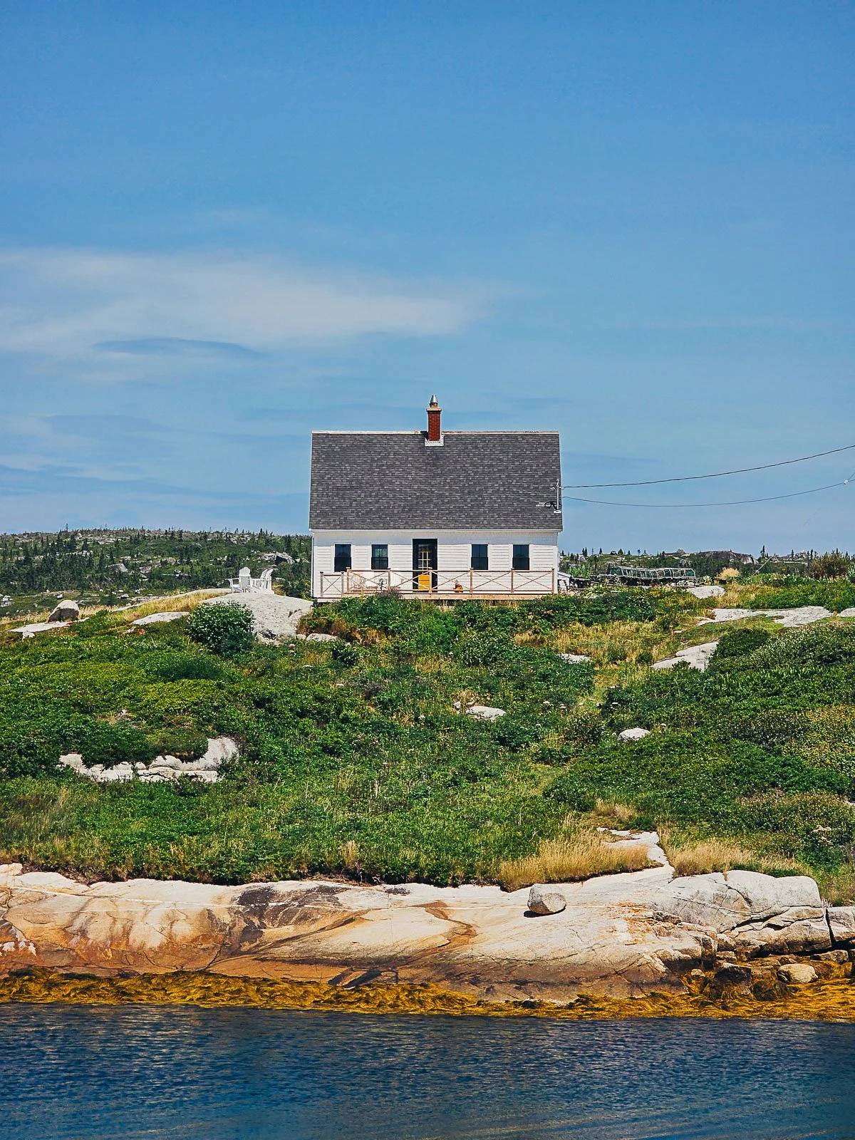 A single white house with a yellow door on the top of a grassy hill. At the bottom of the hill is water and the skiy is clear and blue