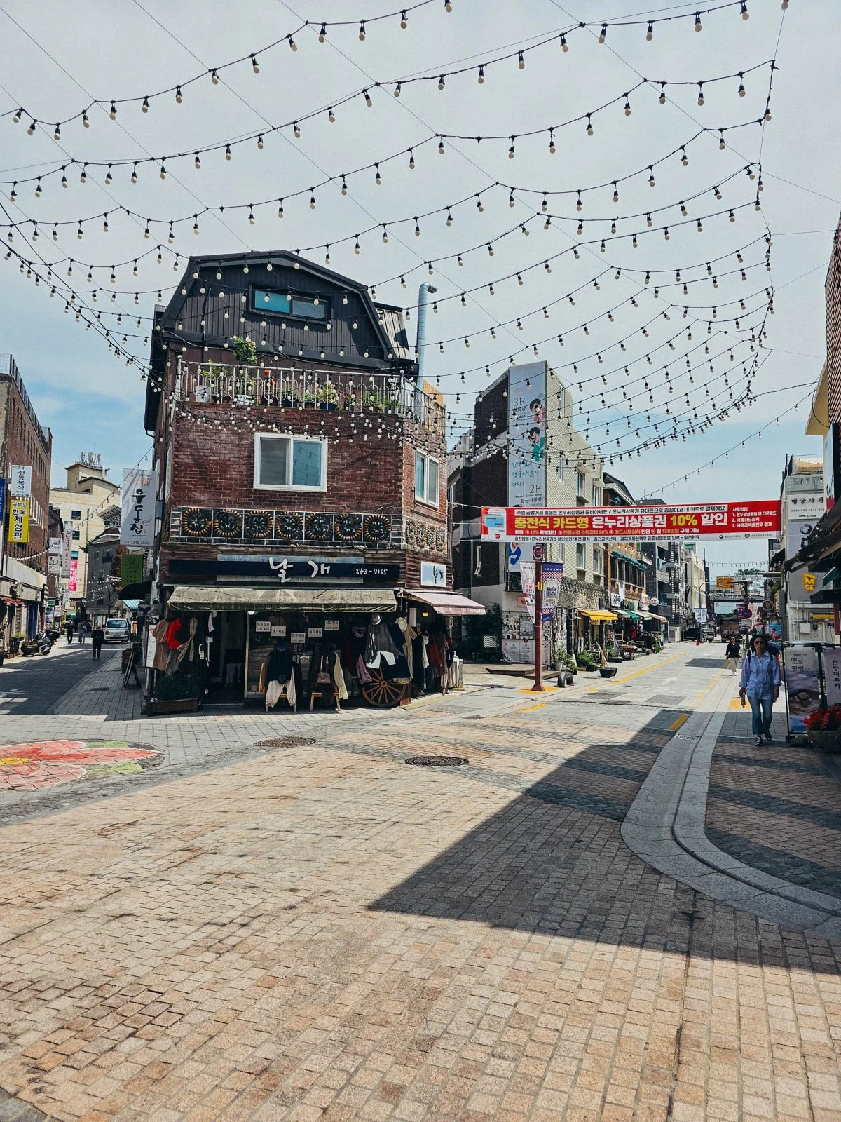 A street with many different shops. A large array of decorative string lighting hang over the street