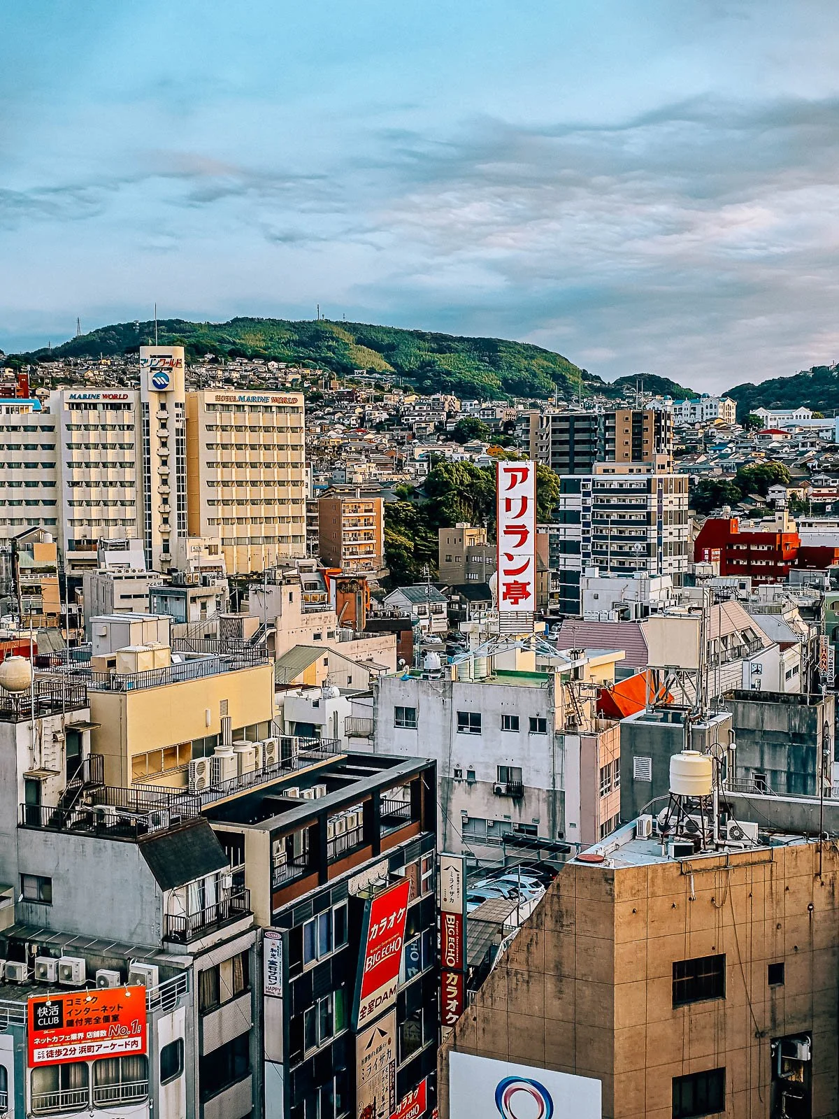 A view from our hotel room looking out across the patchwork of rooftops and tower blocks in donwtown Nagasaki, a green tree covered hill in the distance