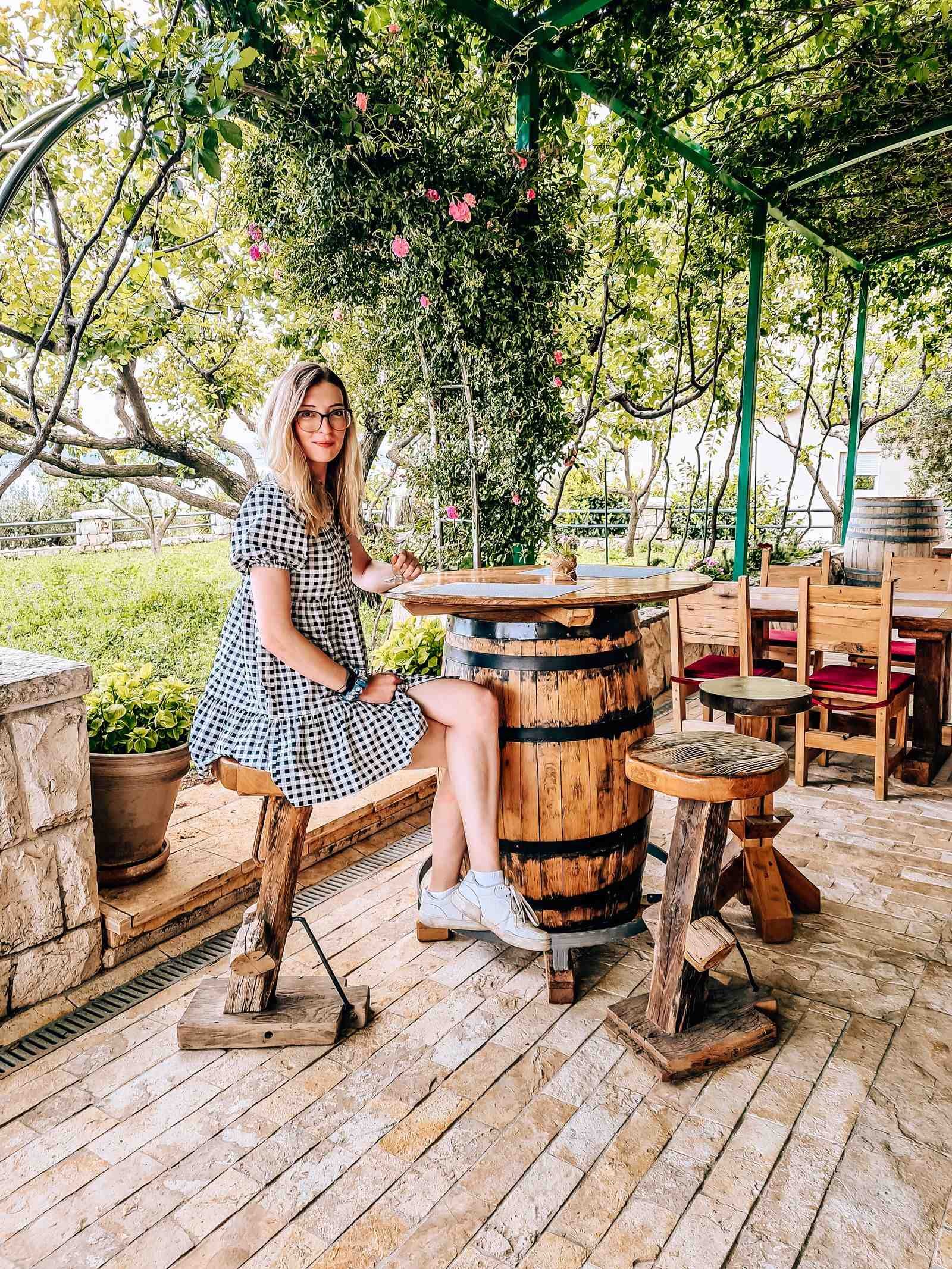 A girl in a black and white dress sitting on a wooden stool leaning against an wooden barrel table underneath many green vines shading the area