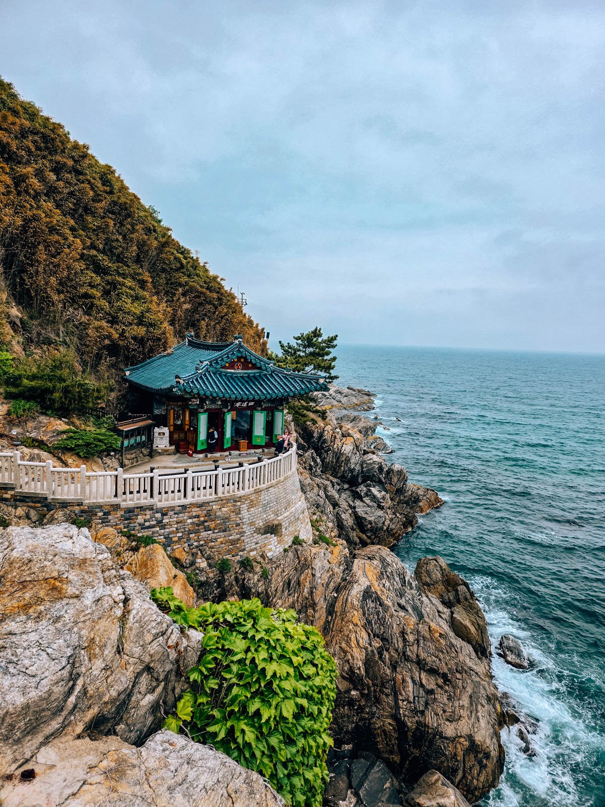 A temple tucked into the side of a rocky cliff face over the sea with a narrow white path leading to the front of the temple