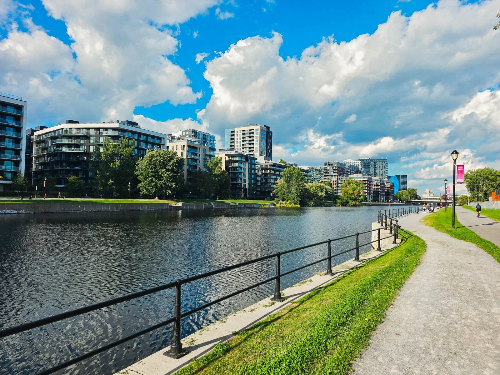 A path runs alongside a canal with a city skyline visible beyond on a blue sky sunny day, one of the best things to do in Montreal on your Canada itinerary