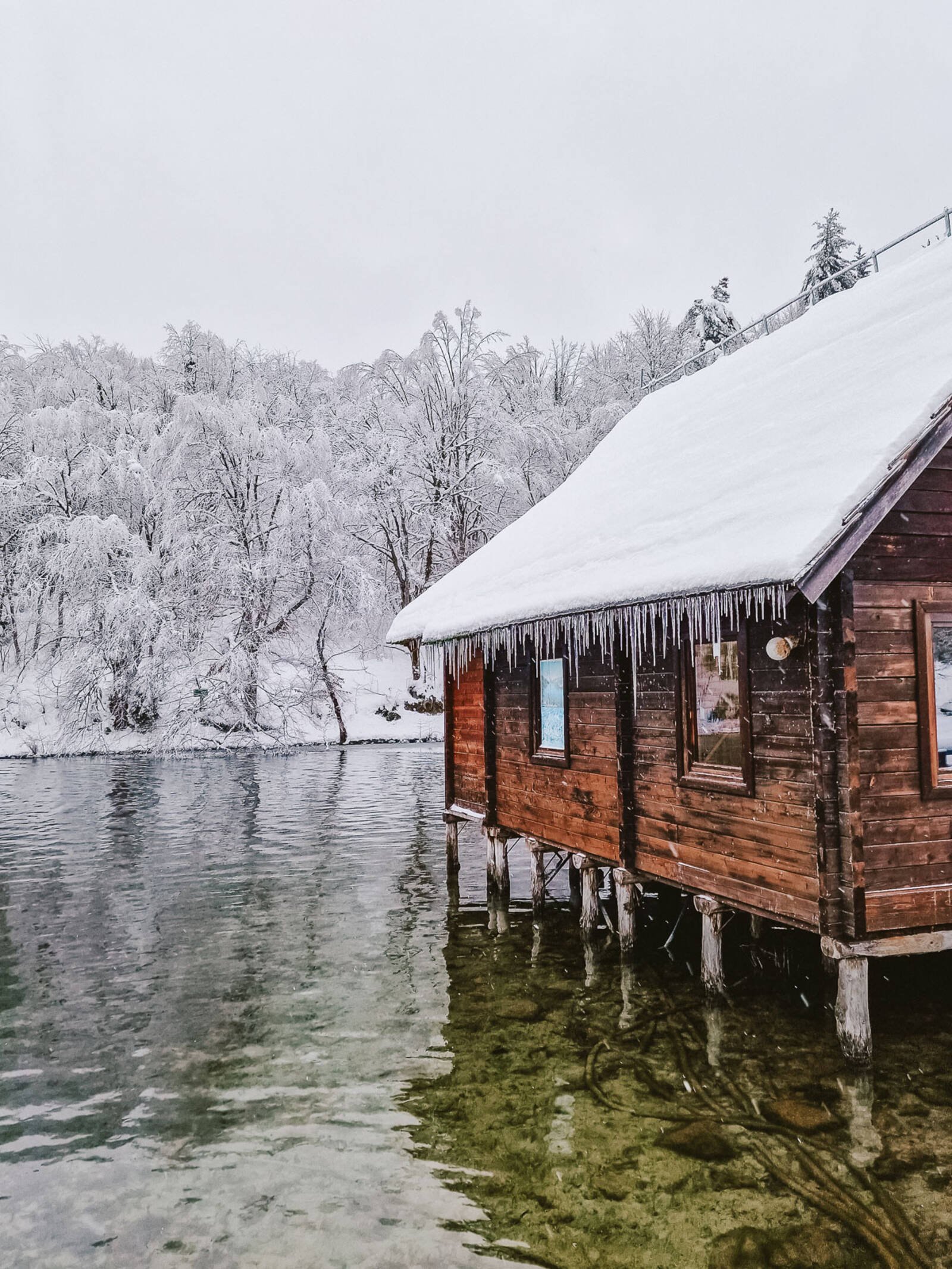 A wooden boat house on stilts above the lake with a snow covered roof and icicles hanging off the roof with trees on the water bank in the background