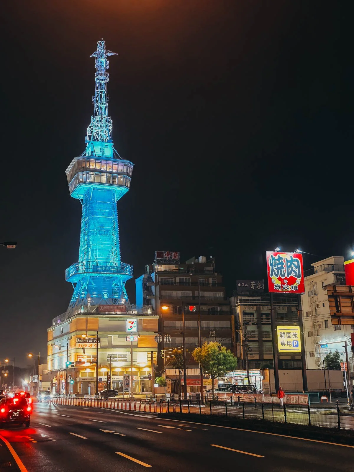 A large tower lit up blue along a street with many shops and buildings at night