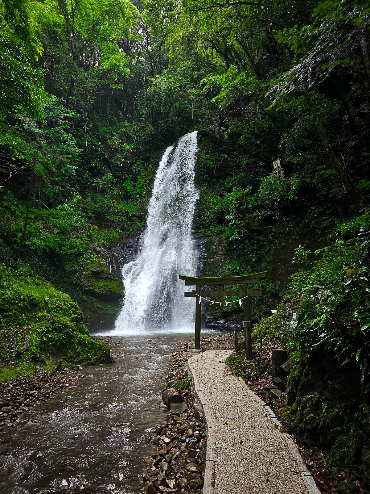 Looking down a stone path running a lot a small river that leads to a single torii gate framing a roaring waterfall from the high surrounding lush green cliffs above