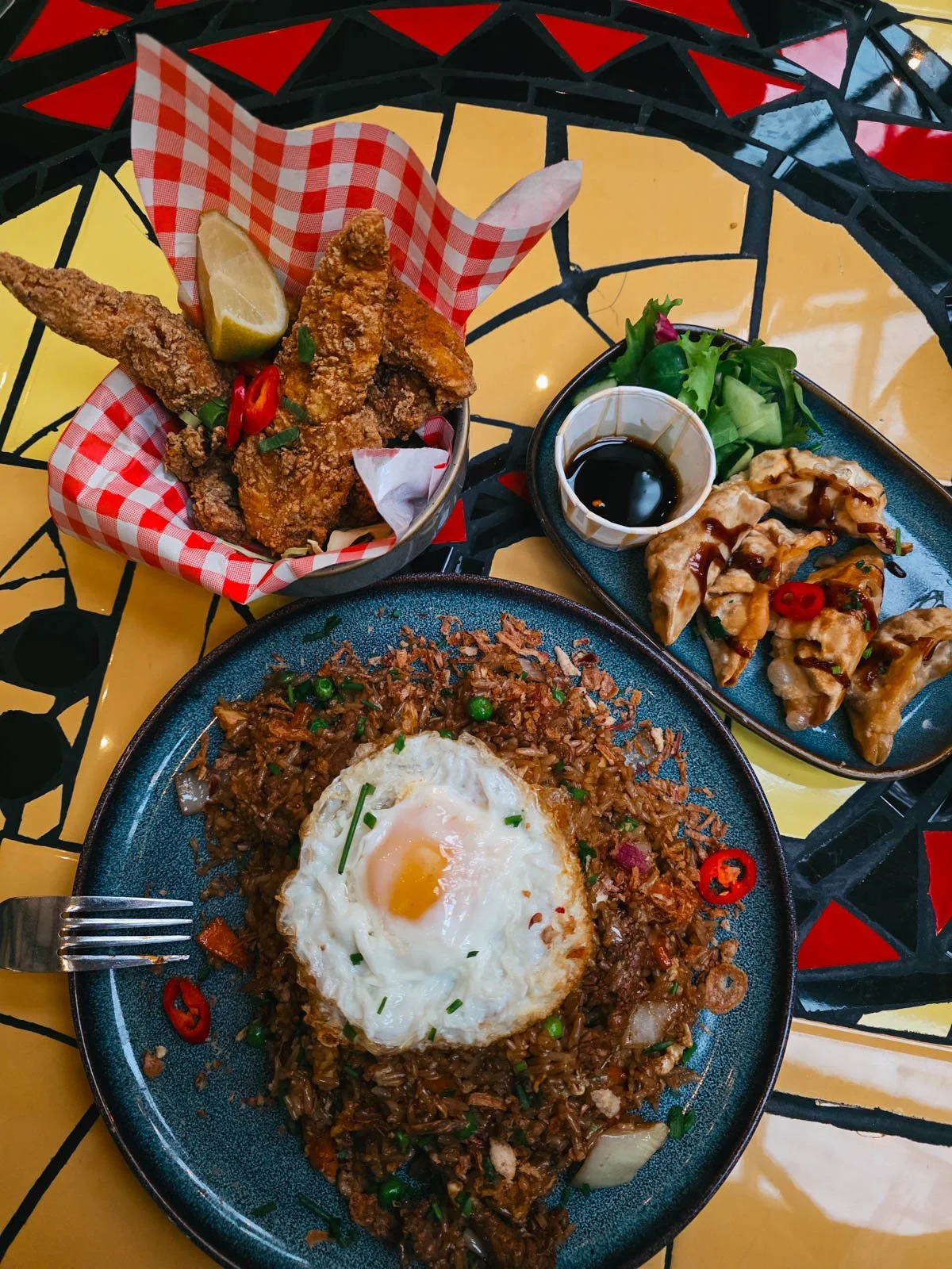 Three plates of food on a yellow tile table, one is fried rice with a fried egg on top, one is a basket of fried chicken, another is gyoza with a soy sauce pot