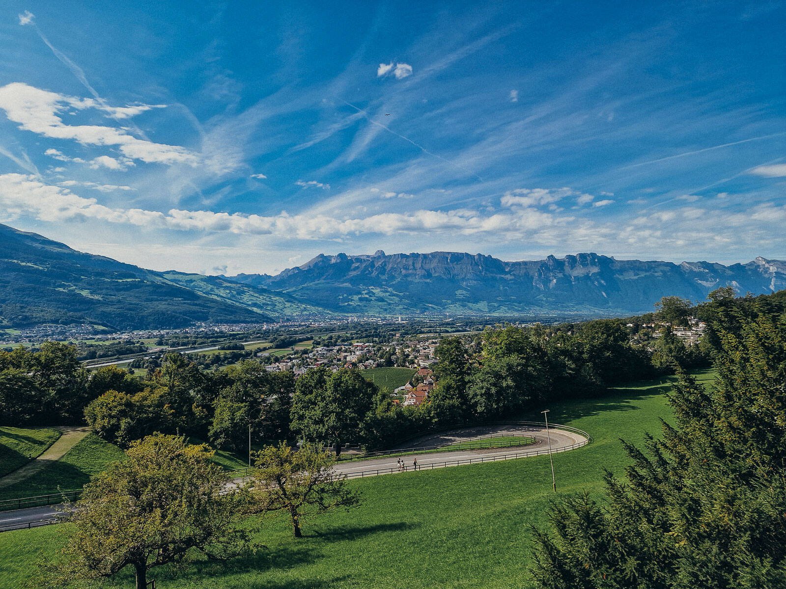 Panoramic image of Liechtenstein with lush green grass in the foreground, trees and a town in the distance with large mountains behind and blue sky