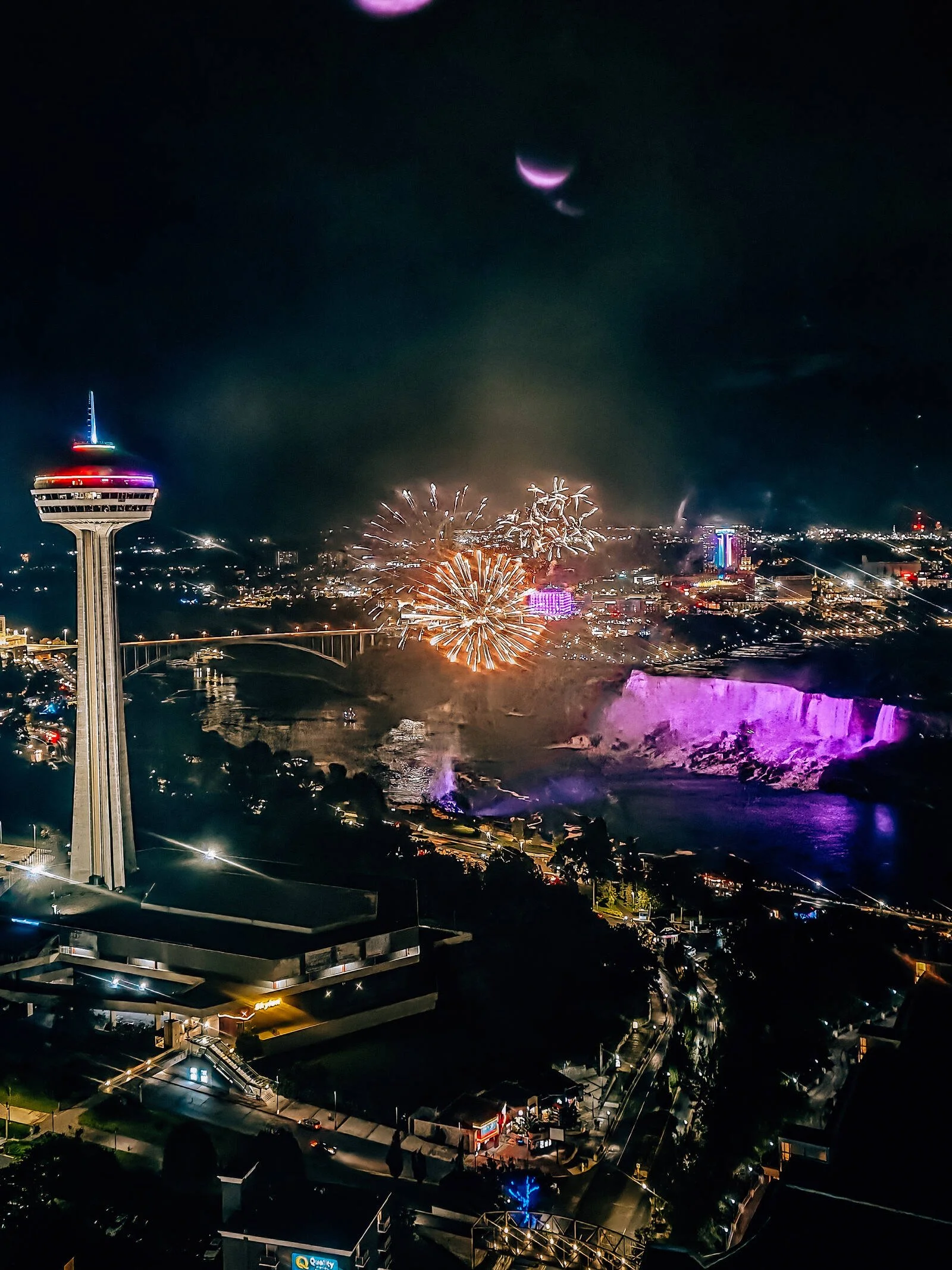 A nighttime view from above of a citty lit up at night, a large viewing tower and fireworks over a river at Niagara Falls, Canada