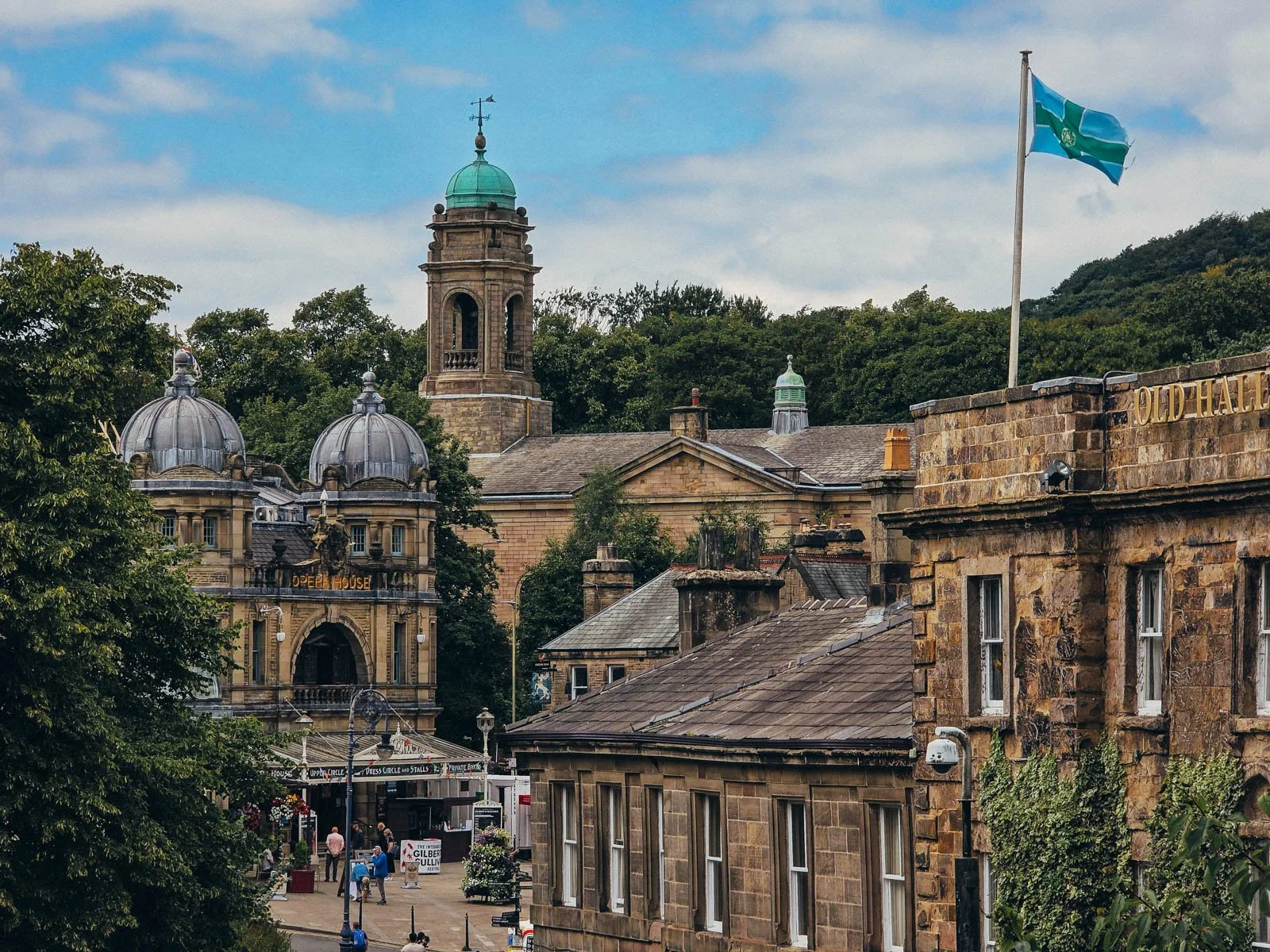Layers of sand-coloured stone buildings in the spa town of Buxton in the Peak District, the corner of a hotel on the right, the front of the opera house with two domes on the left and a spire of a church behind