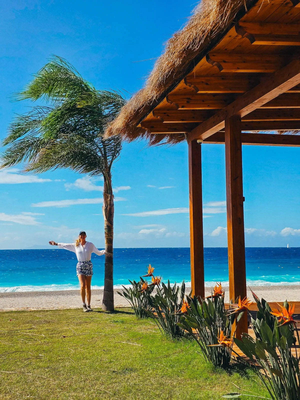 Helena standing next to a palm tree witha a large wooden beach shelter with a sandy beach and blue water in the background