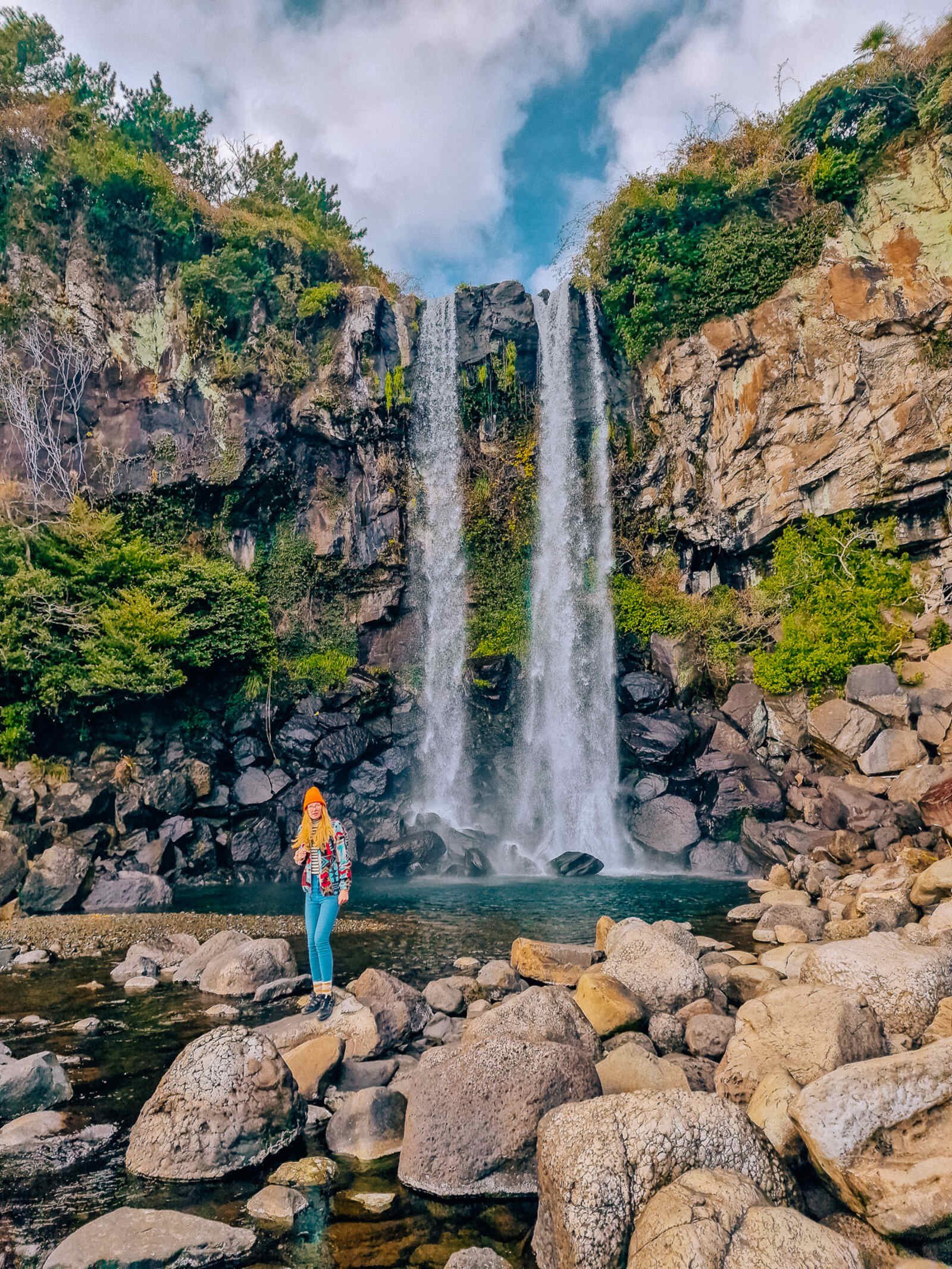 a woman standing on a boulder surrounded by shallow water, in front of a large waterfall cascading down a green moss covered cliff into a dark blue pool