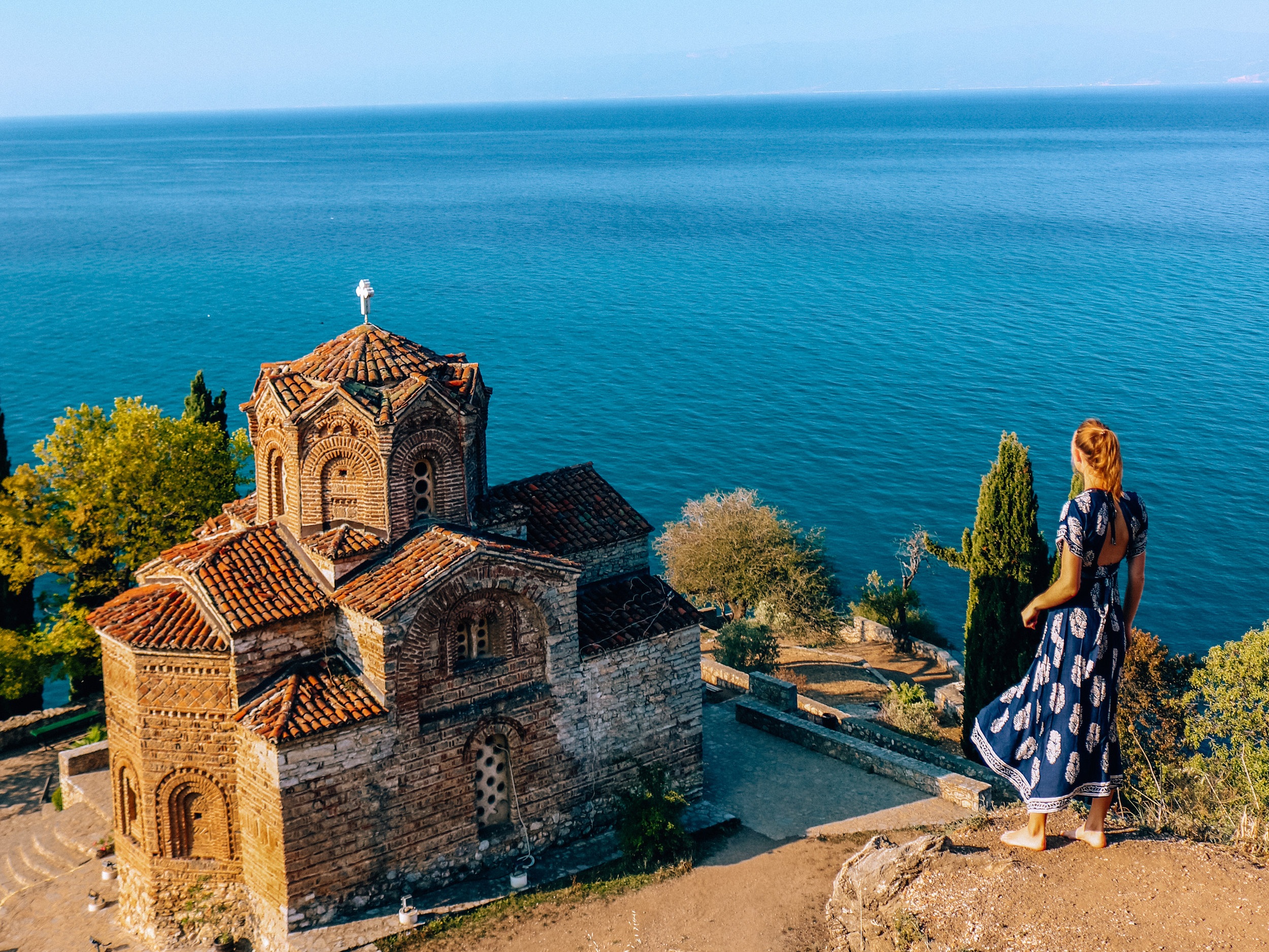Girl in a blue dress standing on the edge of a ridge looking down at an old stone church with the blue water of Lake Ohrid in the distance