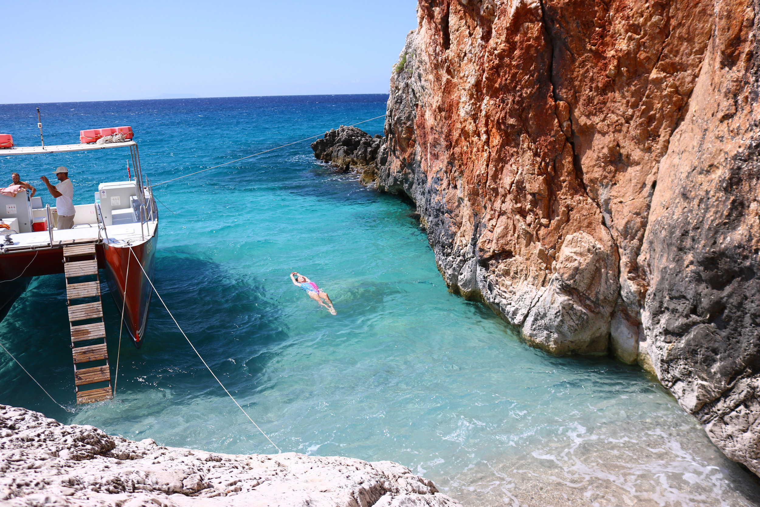 girl floating in turquoise water next to a boat with red cliffs right next to her