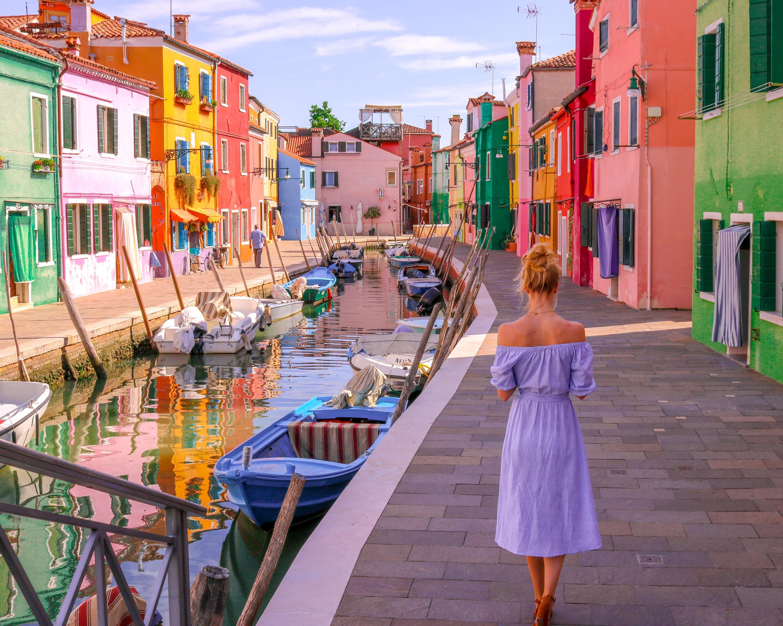 A girl in a blue dress walking along the side o fa pathway with many small boats docked in wather stretching down the middle of the pathway. Each side of the pathway is surrounded by many colourful houses
