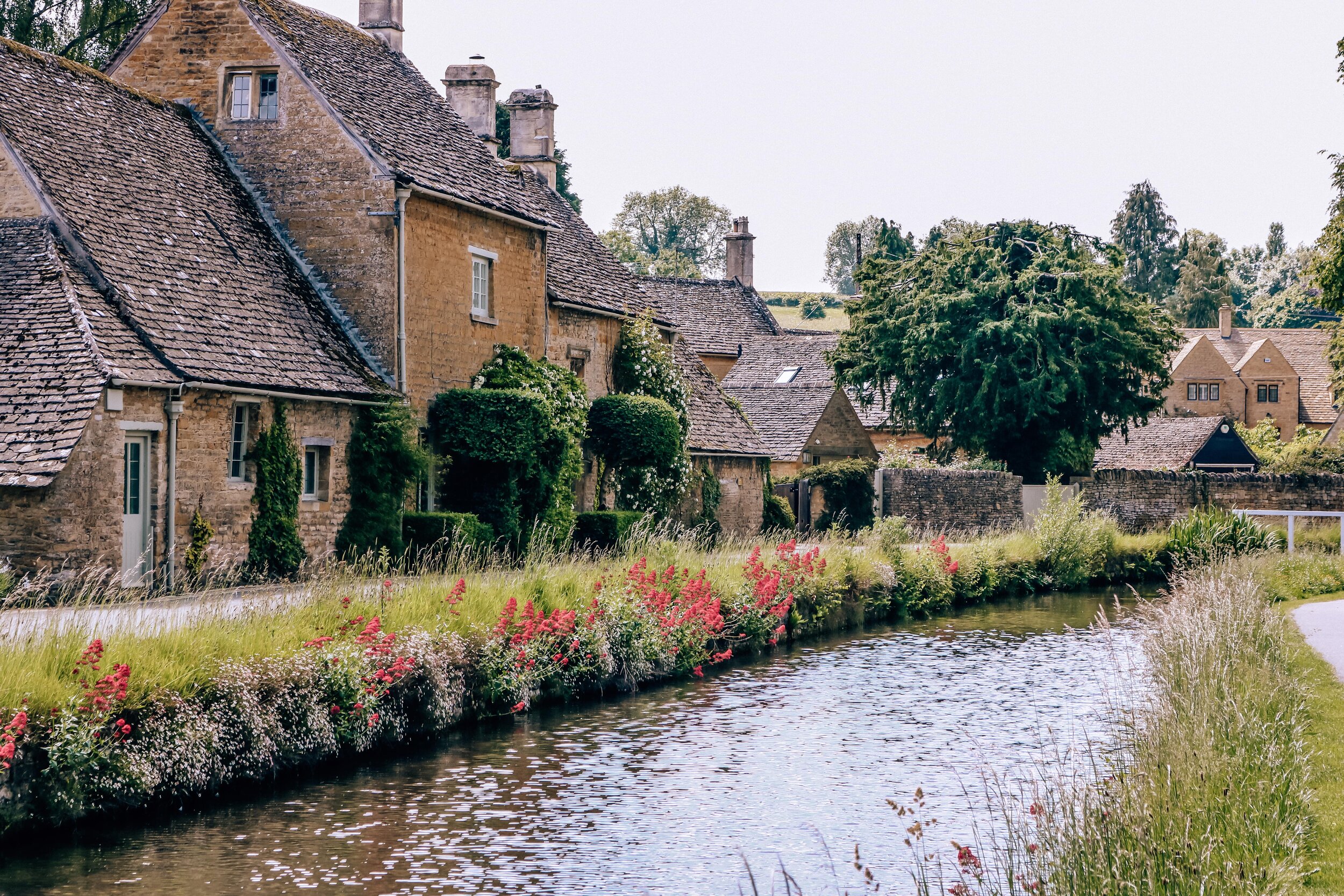 A row of sand-coloured stone houses along a path right next to a stream lined with pink flowers in the Cotswolds