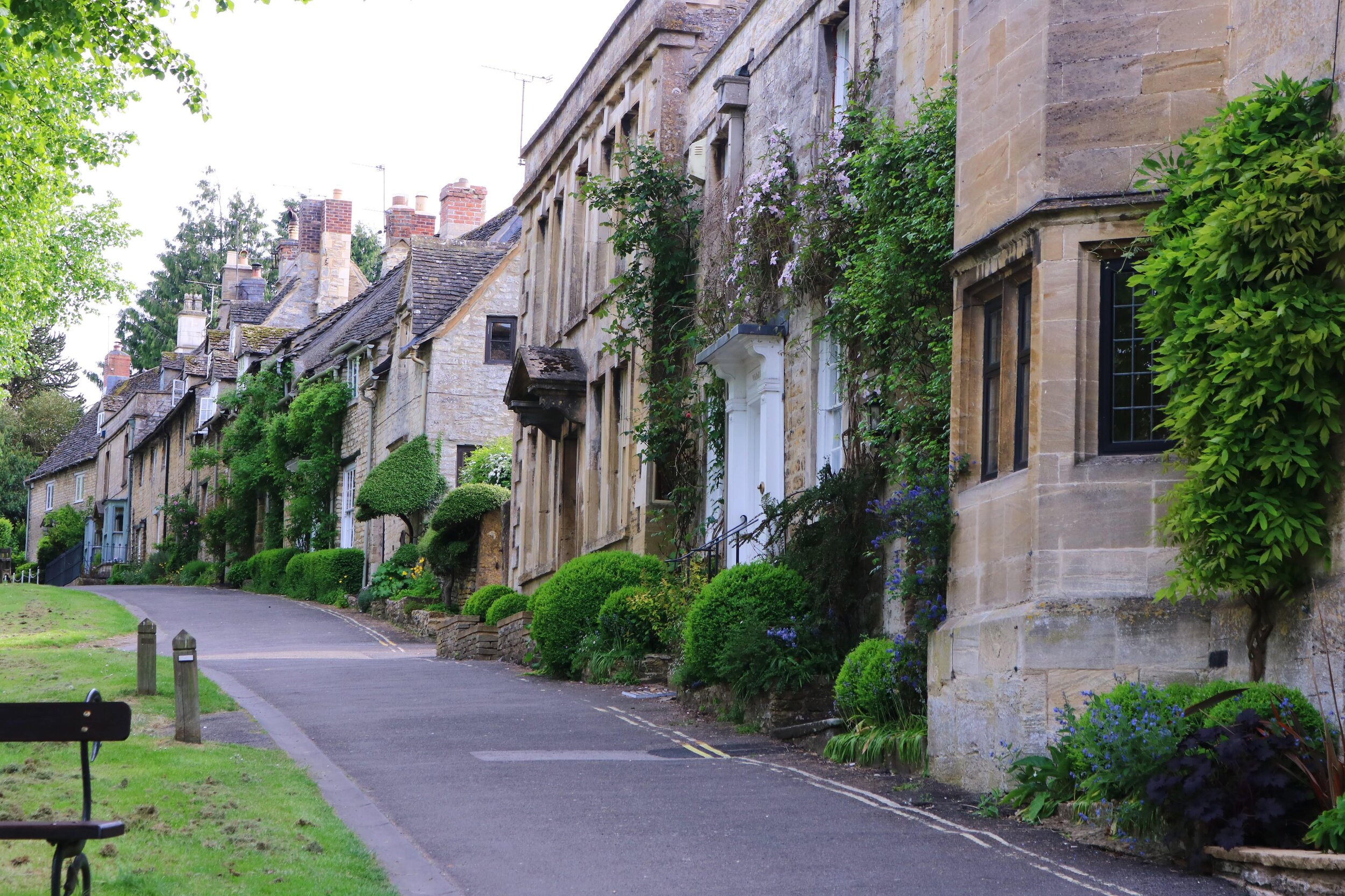 Looking uphill along a path lined with sand-coloured stone houses in the Cotswolds village of Burford with green leafy shrubs outside each front doot