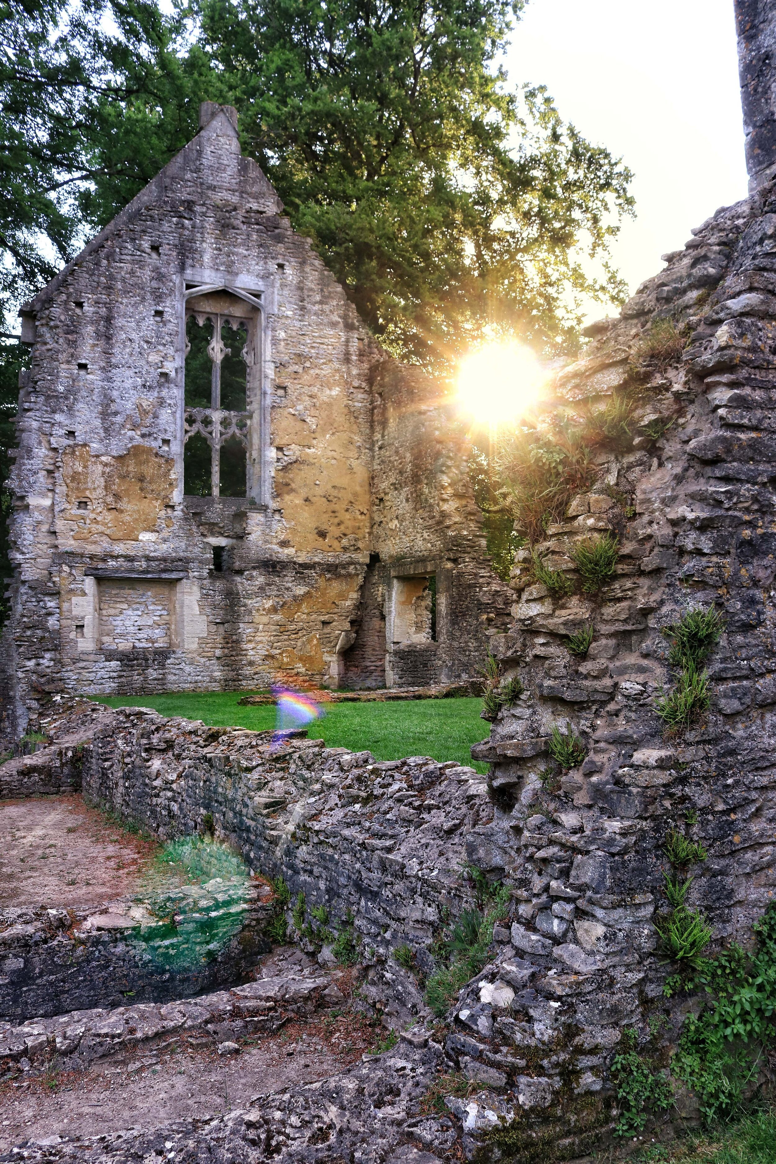 the ruins of an old church in one of the villages in the Cotswolds. The sun shines through a tree sitting behind the pointed end of the ruin in Minster Lovell village