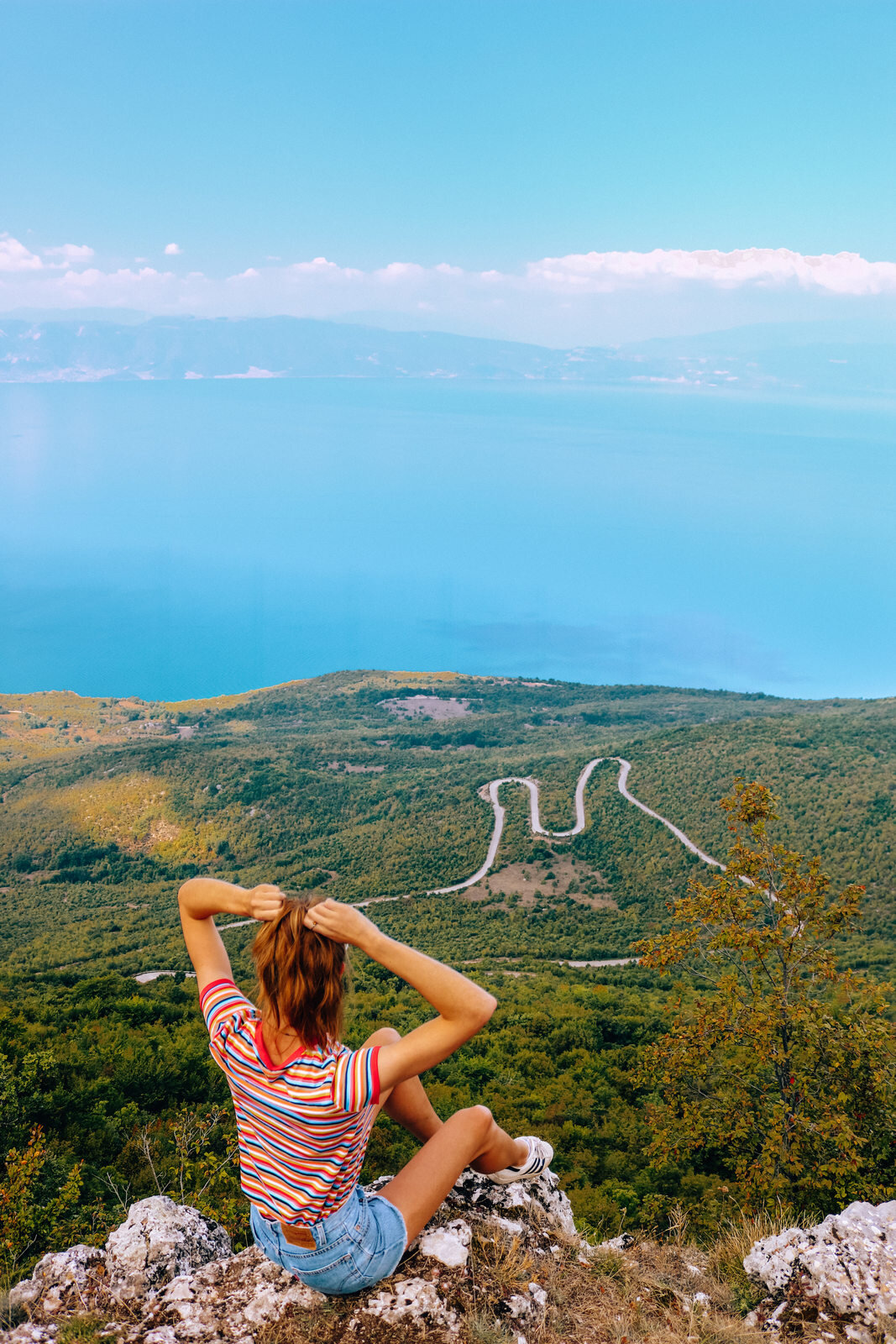 Girl in a colourful strip shirt sitting on a rocky edge looking out onto a windy road snaking through green hills