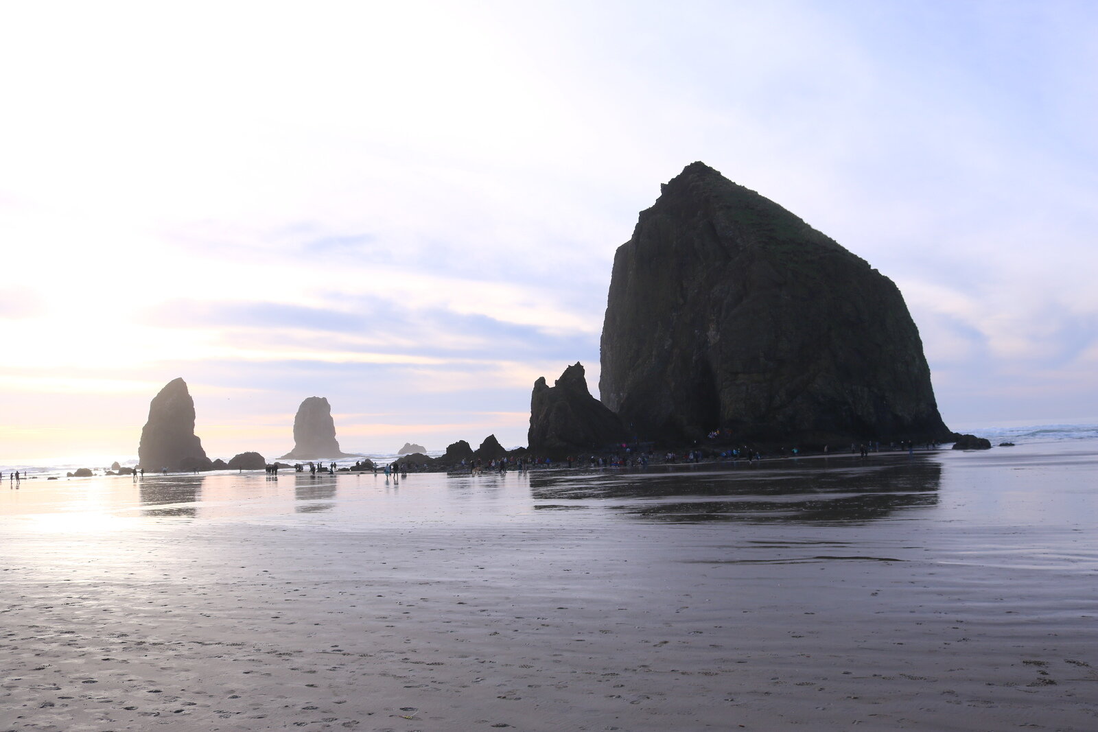 Haystack Rock on Cannon Beach - a large dark rock on the beach at low tide, the ocean is in th distance and the sand is shiny with the sun reflection