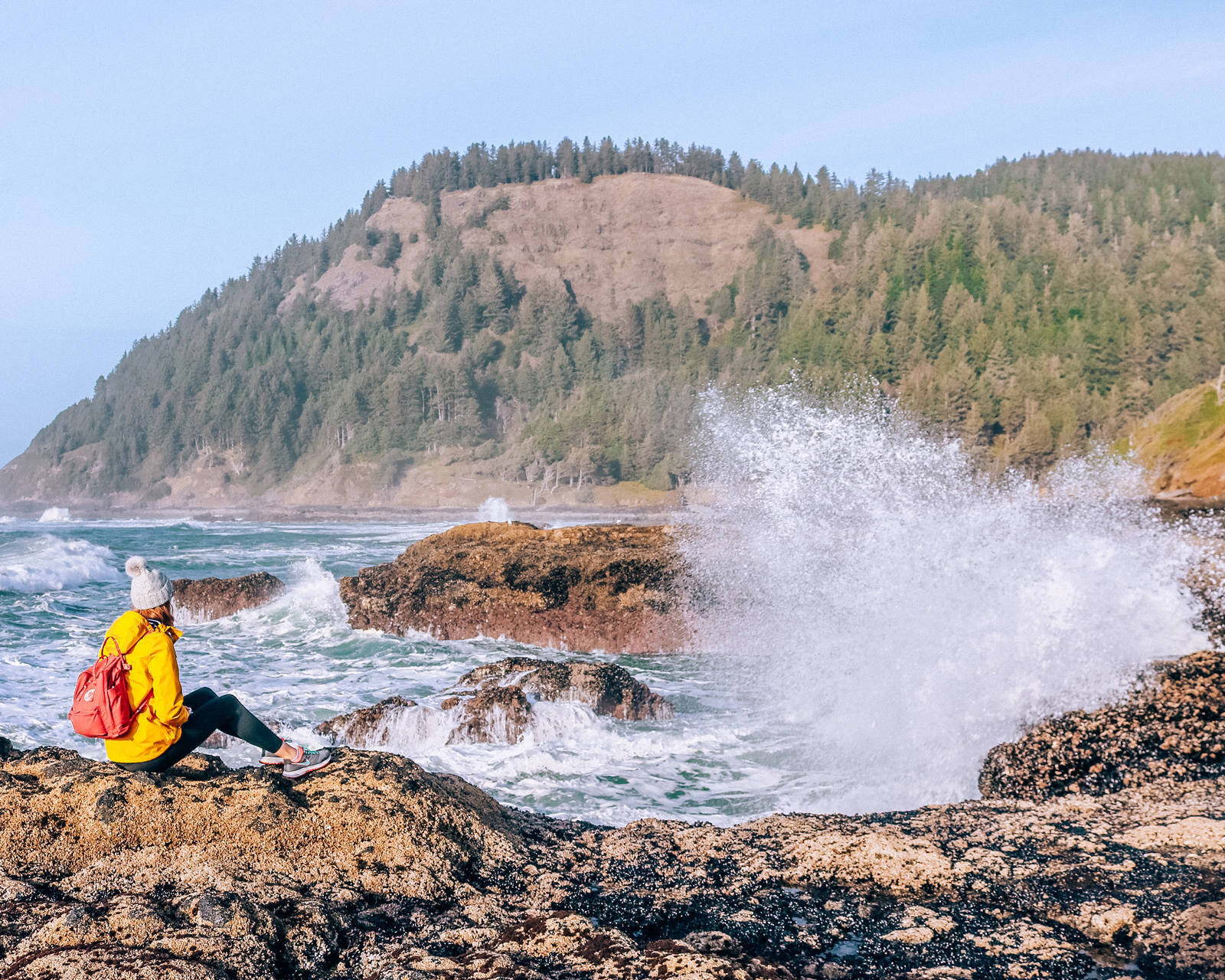 Girl sitting on a rock as a huge wave splashes up and a tree covered hill is in the distance, Thors’s Well, Oregon Coast