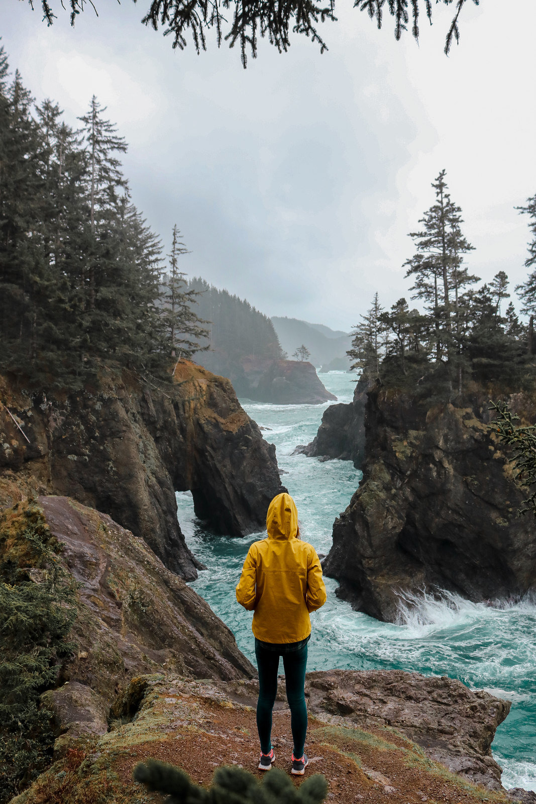 Girl in a yellow jacket looking across a stormy coastal view with turquoise rough ocean waves and dark cliffs covered in mist and green trees,Samuel H Boardman Scenic State Corridor, Oregon Coast