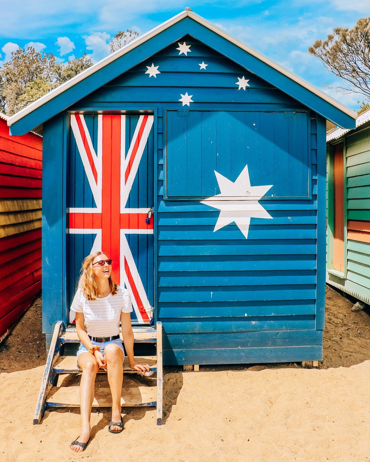 Agirl sitting on the steps of a blue beach hut with the Australian flag on it atBrighton Beach huts in Melbourne, Australia