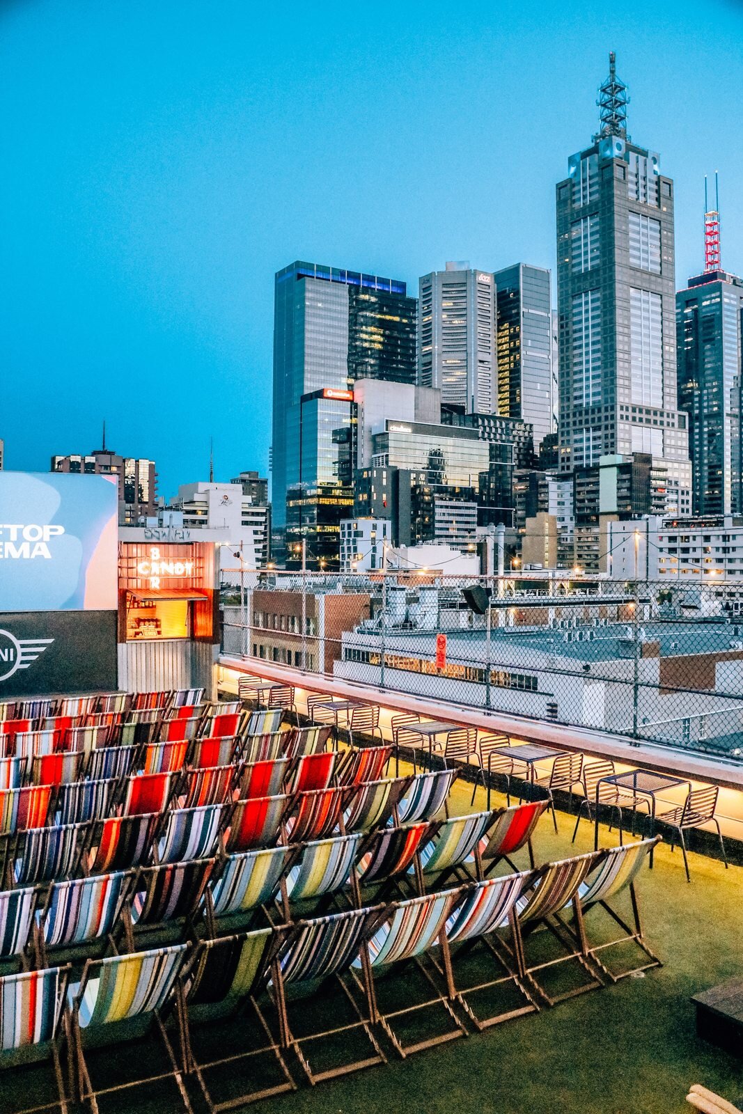 rooftop cinema in Melbourne, Australia at dusk with colourful deck chairs facing a large screen and skyscrapers in the background