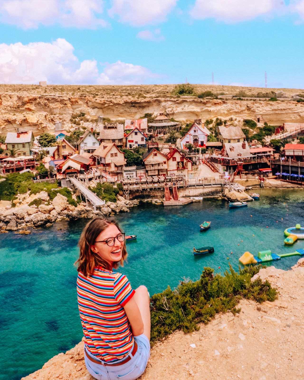 Helena sitting on the edge of a cliff looking out at the blue waters and village of the Popeye Village viewpoint in Malta