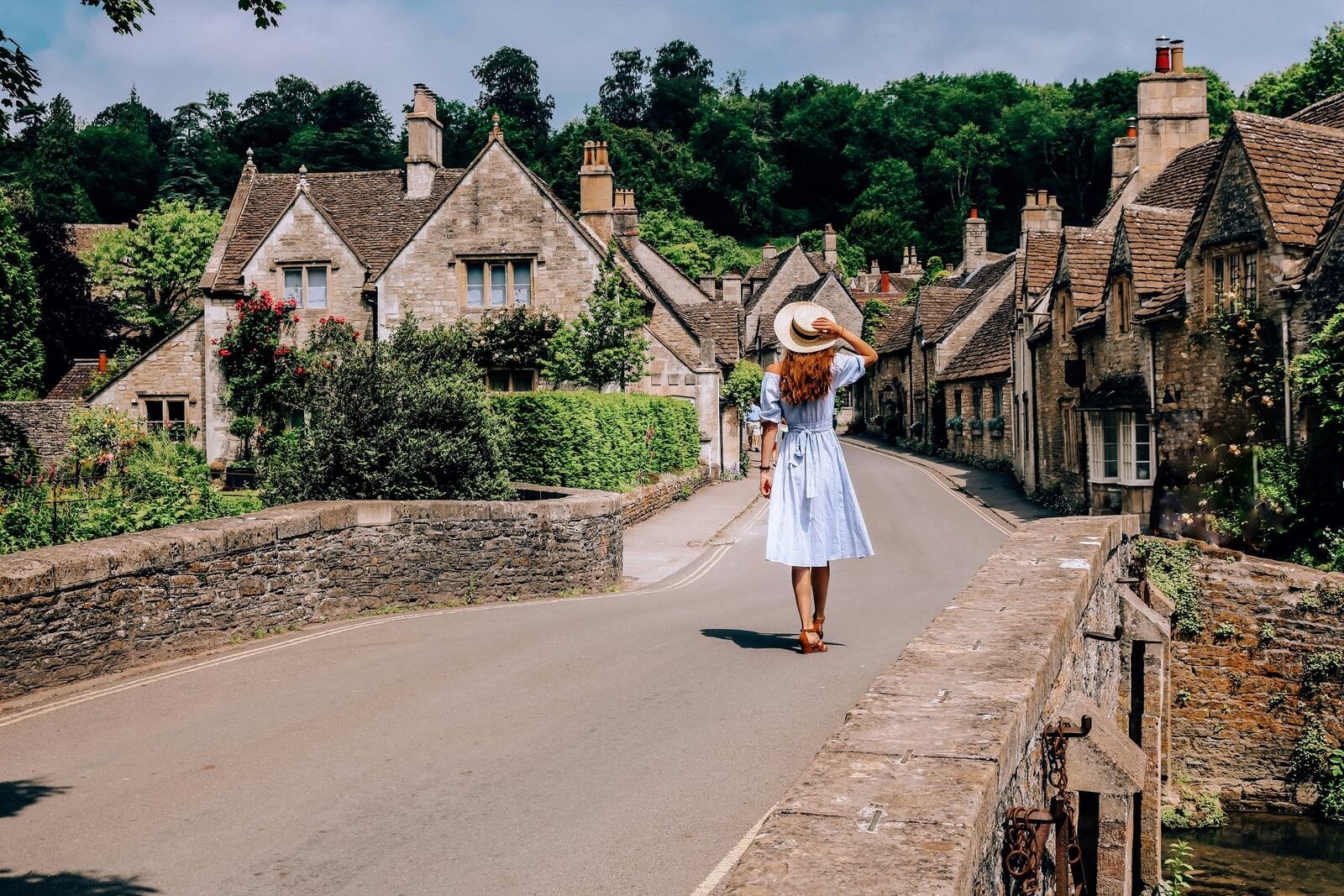 A girl in a blue dress and straw hat walking across a bridge inCastle Combe, The Cotswolds with cute cottages in the distance