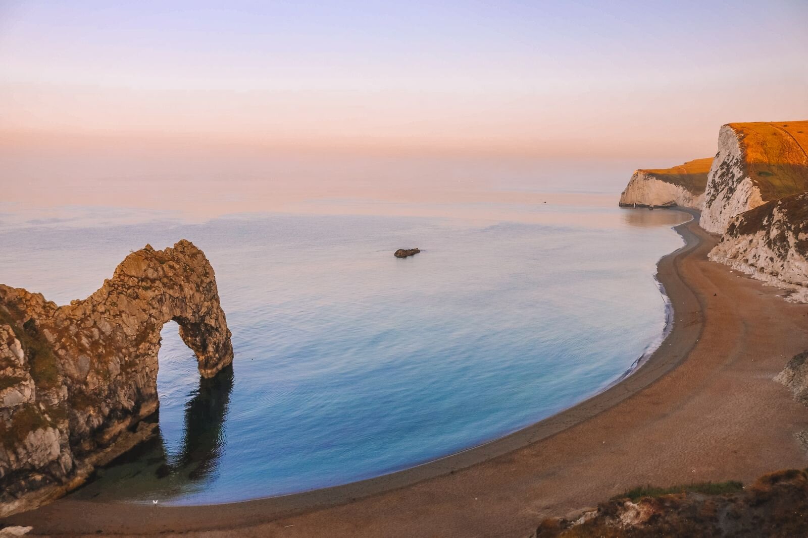 Looking down on a stone archway and beach with white cliffs in the distance at Durdle Door at sunrise