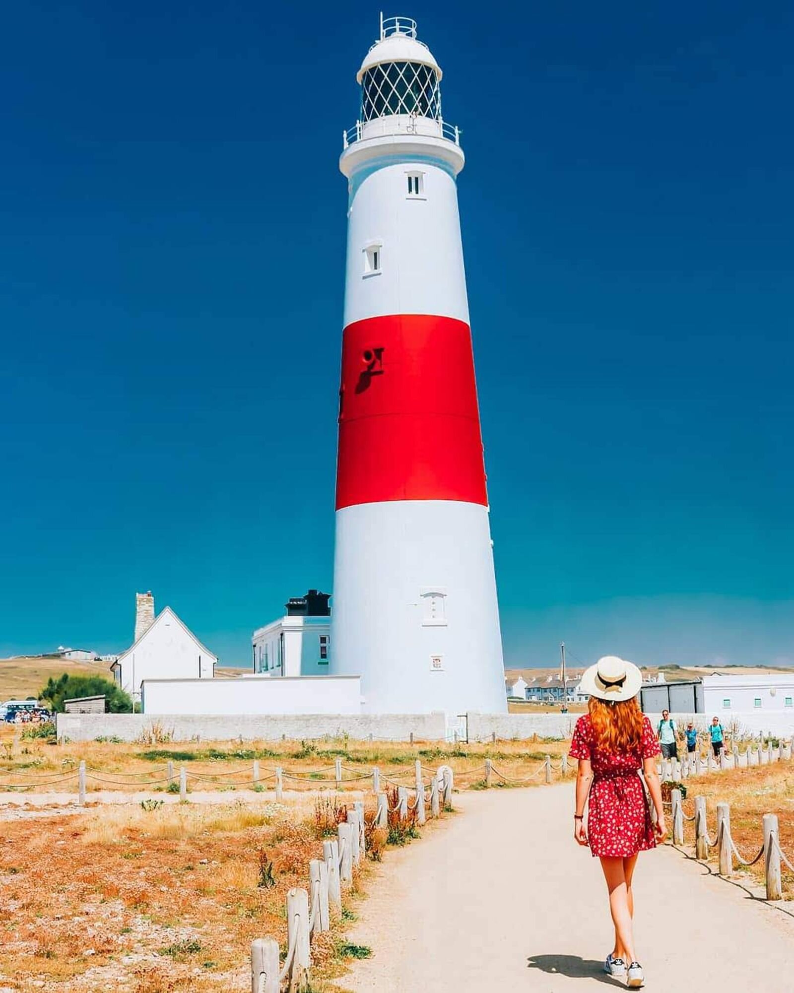 Girl in a red dress walking down a path towards a red and white lighthouse at Portland Bill Lighthouse in Dorset