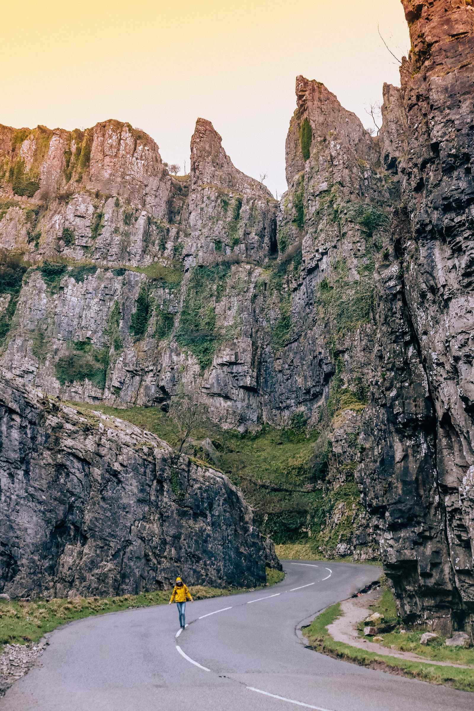 A tiny figure in a yellow coat walking down a winding road in Cheddar Gorge, tall craggy cliffs rise steep on either side