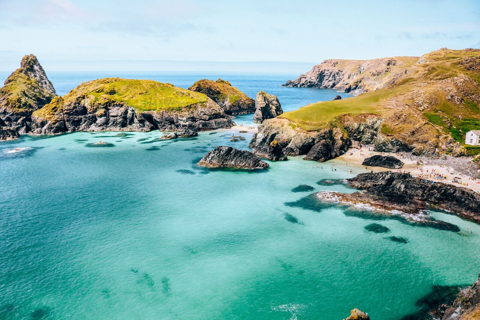 Looking down from cliffs to Kynance Cove, a bay in Cornwall with turquoise blue water, a small beach and lots of rocks protruding from the water with green grassy slopes behind the beac