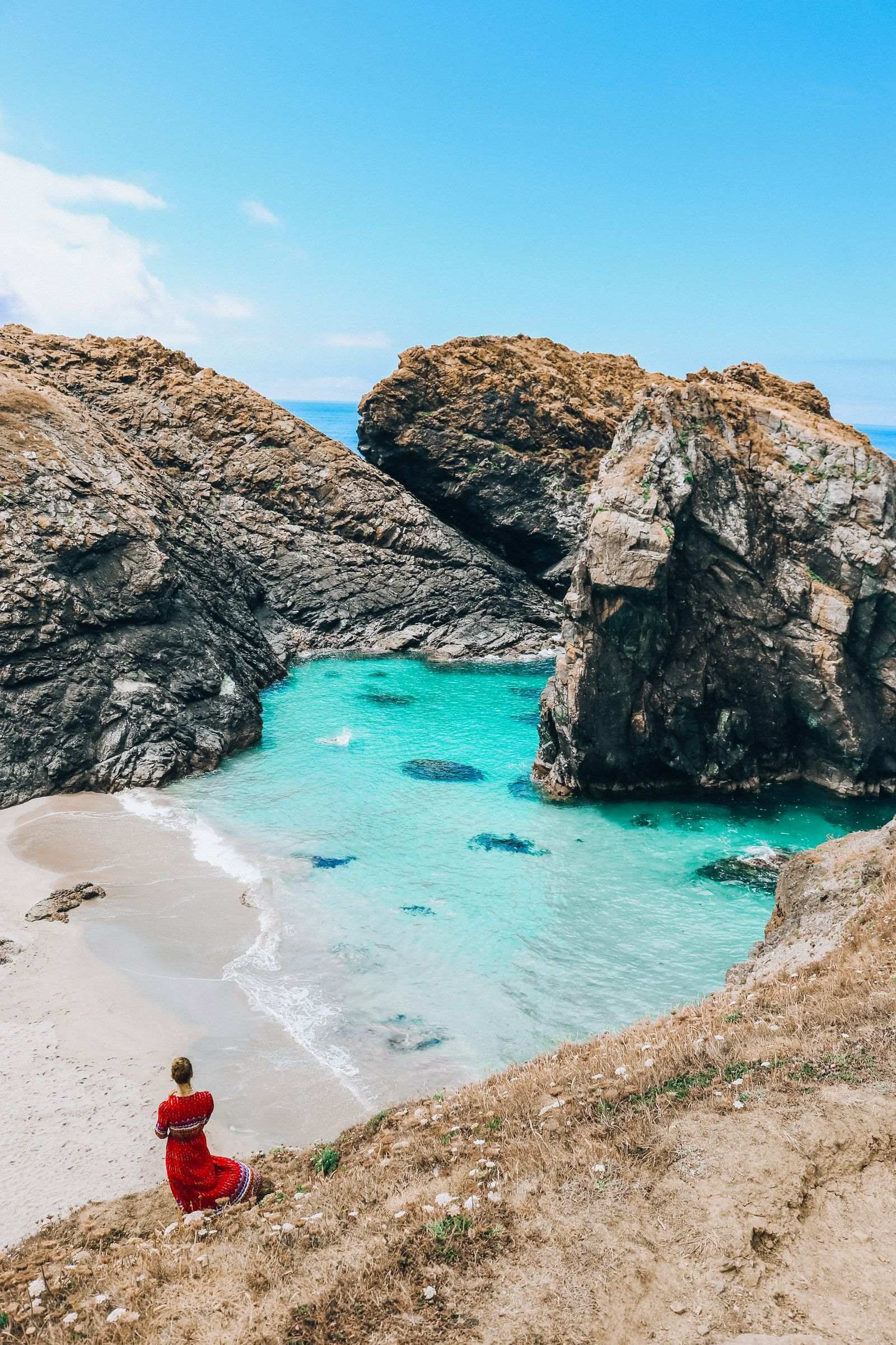 Girl in a red dress standing on a grassy slope with a white sand beach below and turquose blue water lapping at the beach with grey craggy rocks around the cove