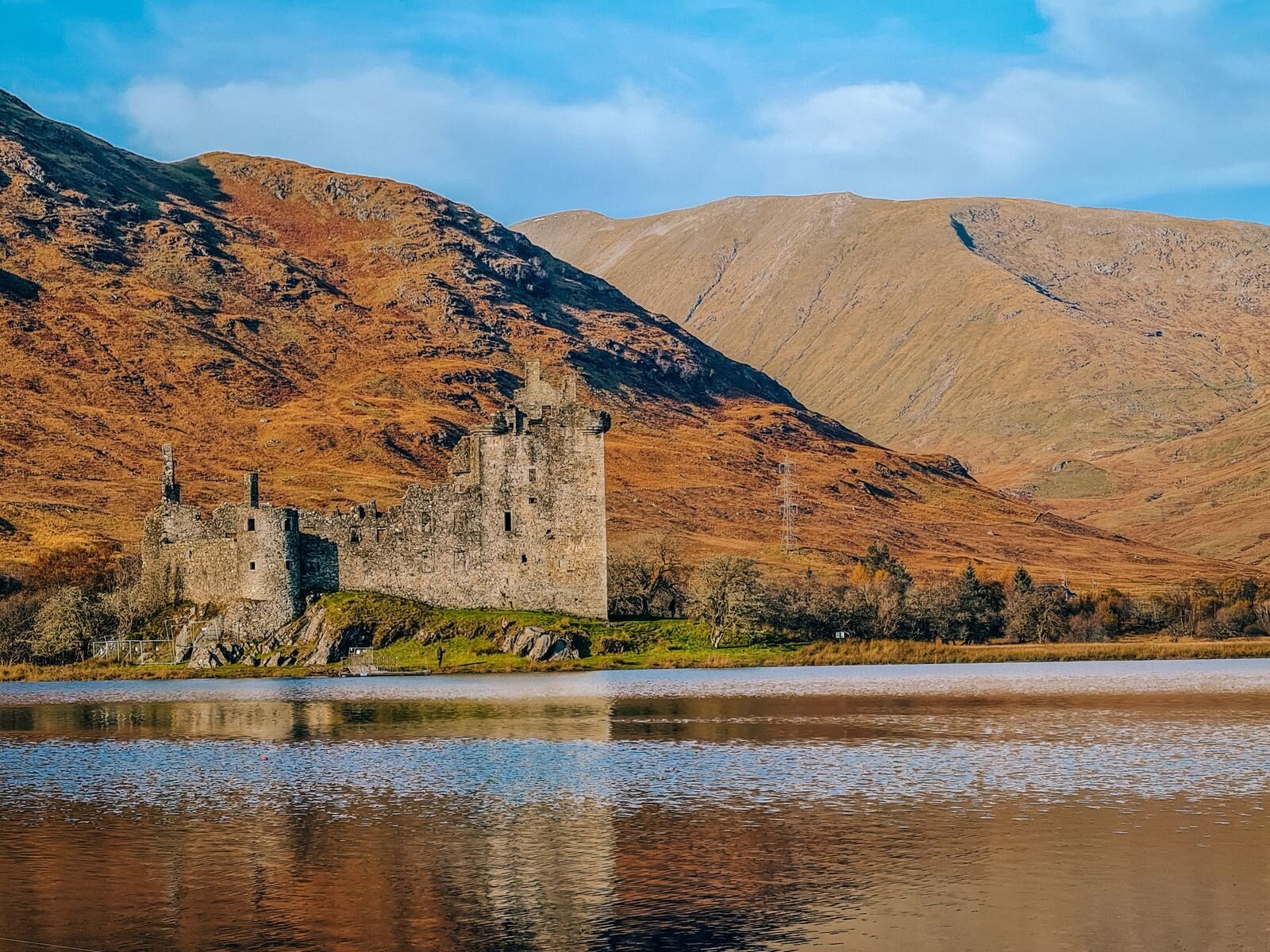 Looking across a lake with castle ruins on the far shore and mountains behind, covered in orange heather. Kilchurn Castle in Scotland