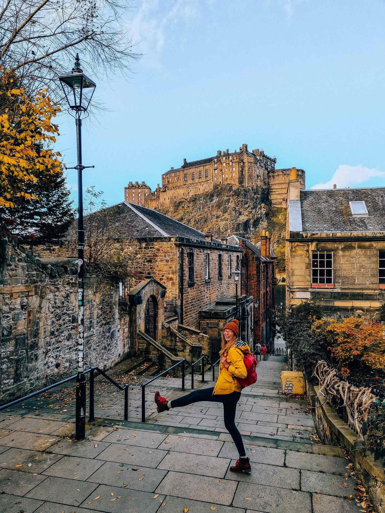 A girl on the stone steps at the Vessel viewpoint of Edinburgh Castle, she wears a yello coat and a wooly hat and is kicking her leg out in the air