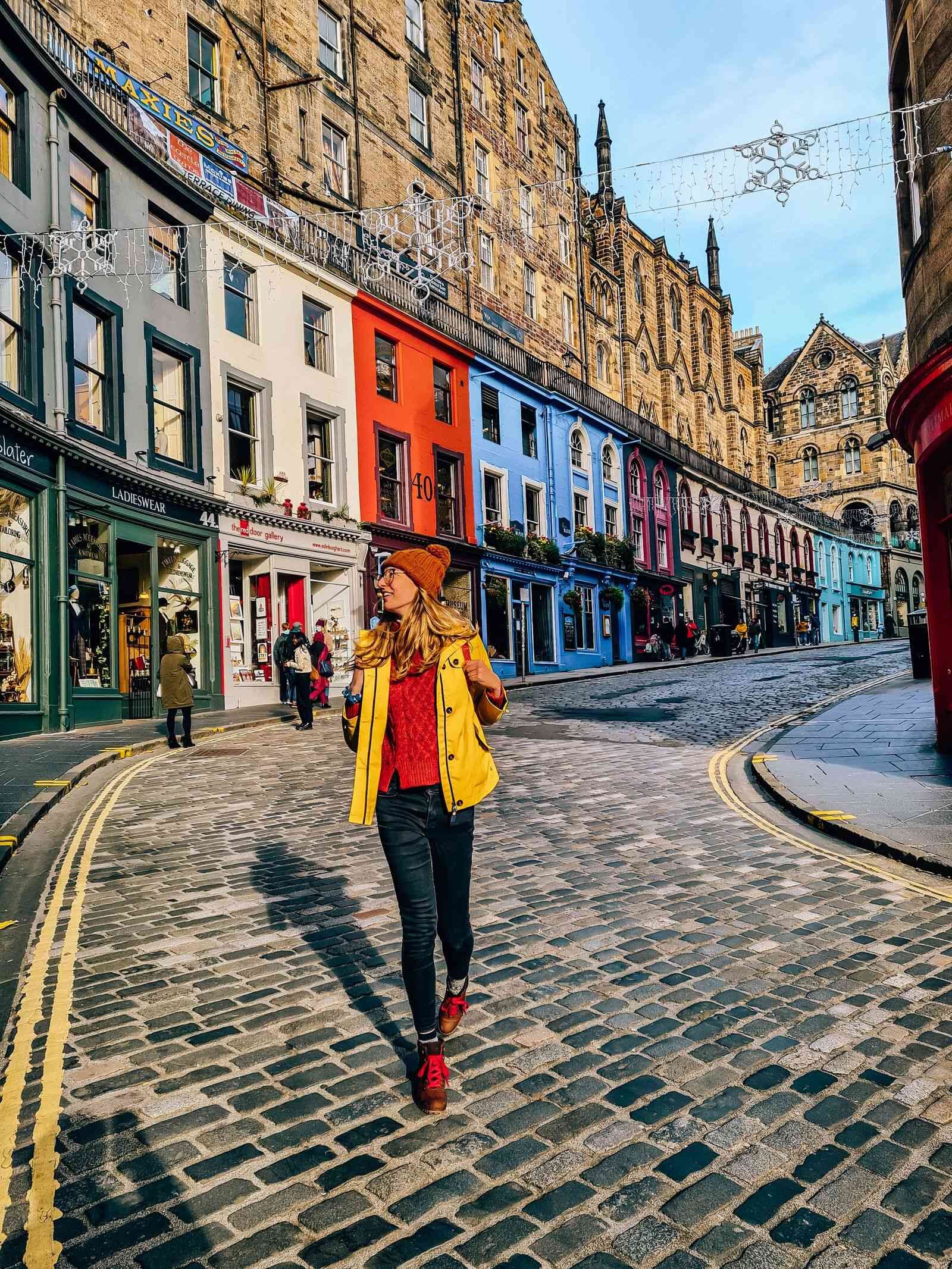 Woman in black jeans, red jumper, yellow coat and woolly hat walking down a cobbled street - Victoria Street, Edinburgh