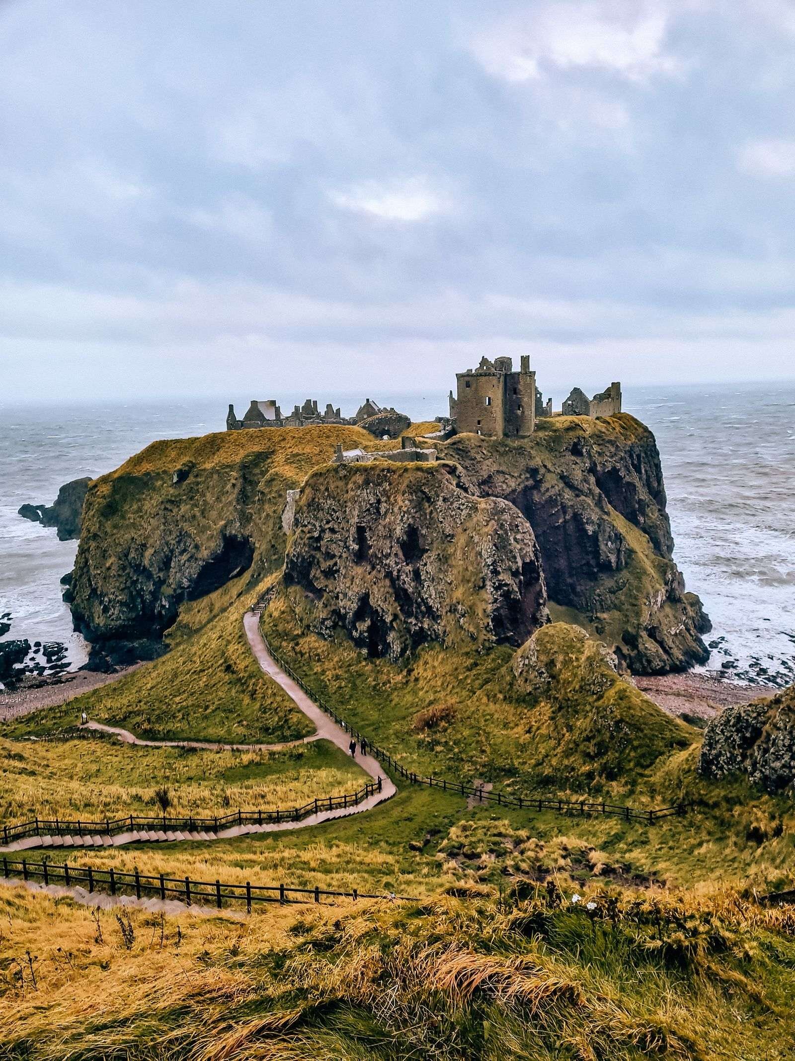 Dunnottar Castle ruins on a rocky outcrop surrounded by grey sea with a single path on a rocky, grassy slope leading to the castle