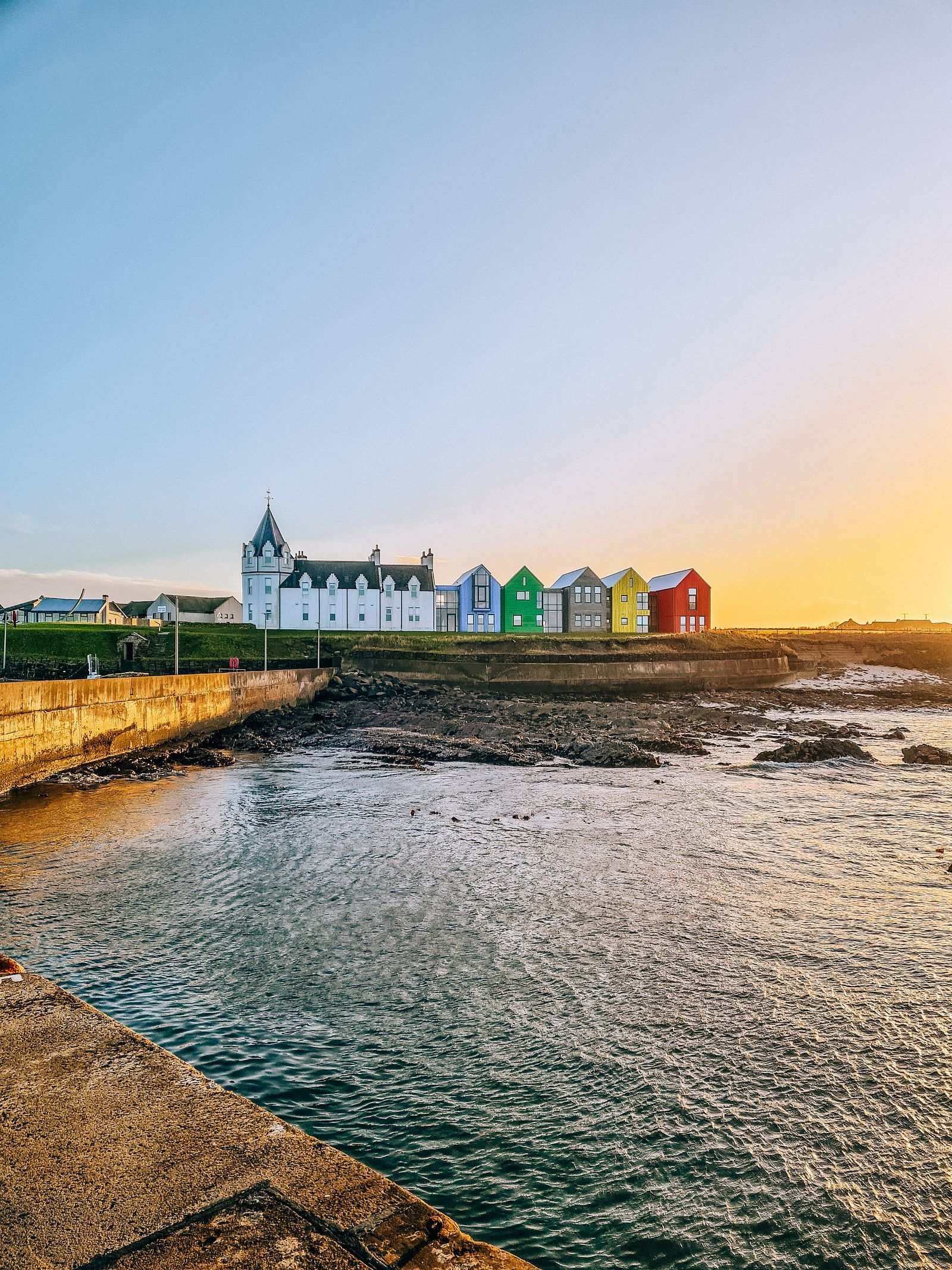A view of John O’Groats Inn from the sea wall, looking back to the land across a harbour. On the land is a white turreted building with 5 colourful buildings next to it in blue, green, grey, yellow and red. Sky is orange at sunset