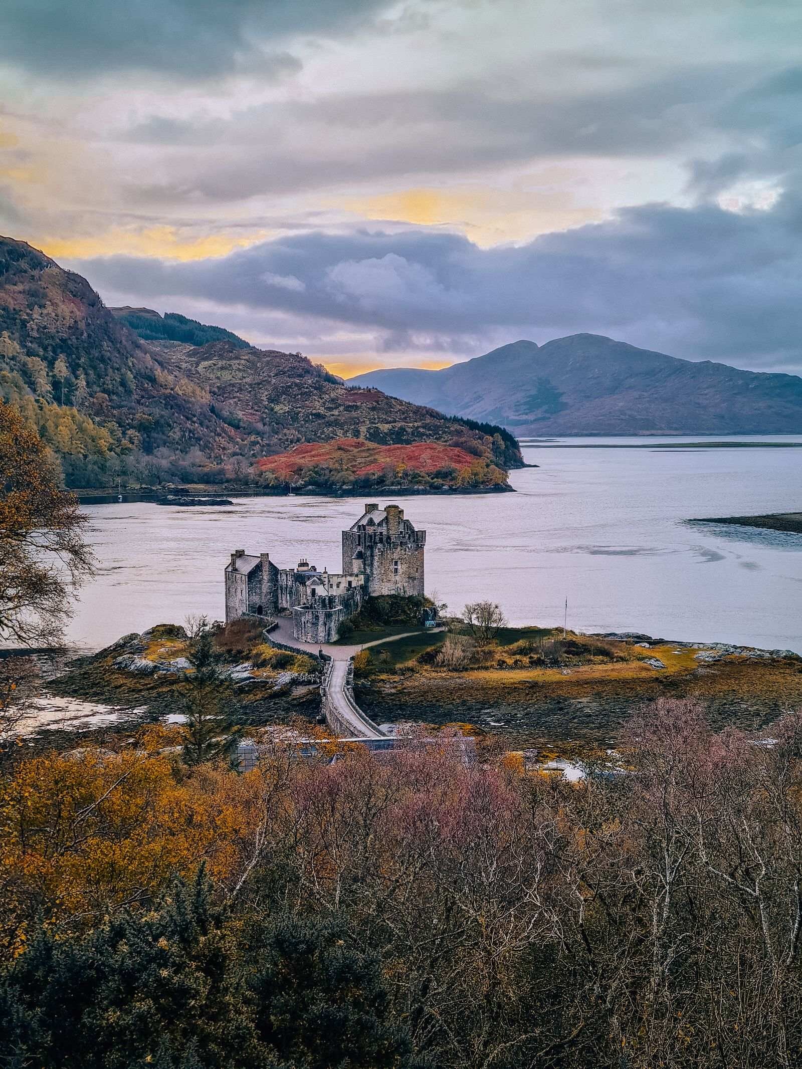 A panoramic view of Eilean Donan Castle - a grey castle on a small island in the lock connected to the mainland by a causeway. Dramatic mountains line the loch behind with low clouds